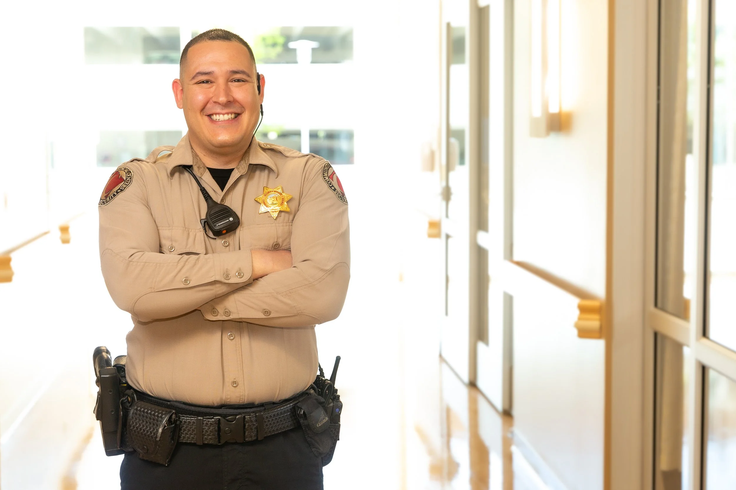 A smiling police officer in tan uniform standing in a well-lit hallway with arms crossed.