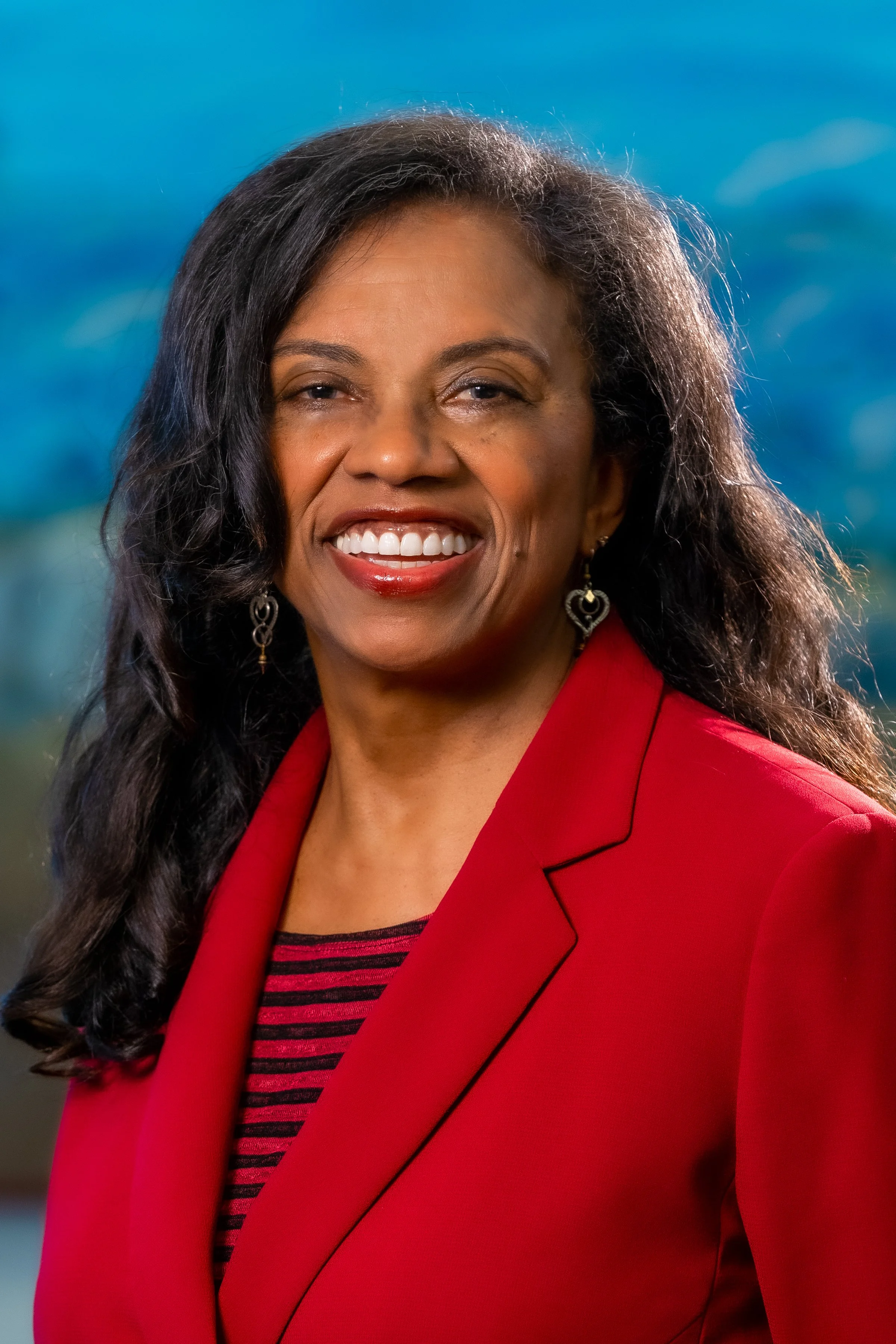 A woman with long black hair, wearing a red blazer and earrings, smiling, with a scenic background of blue sky and green hills.