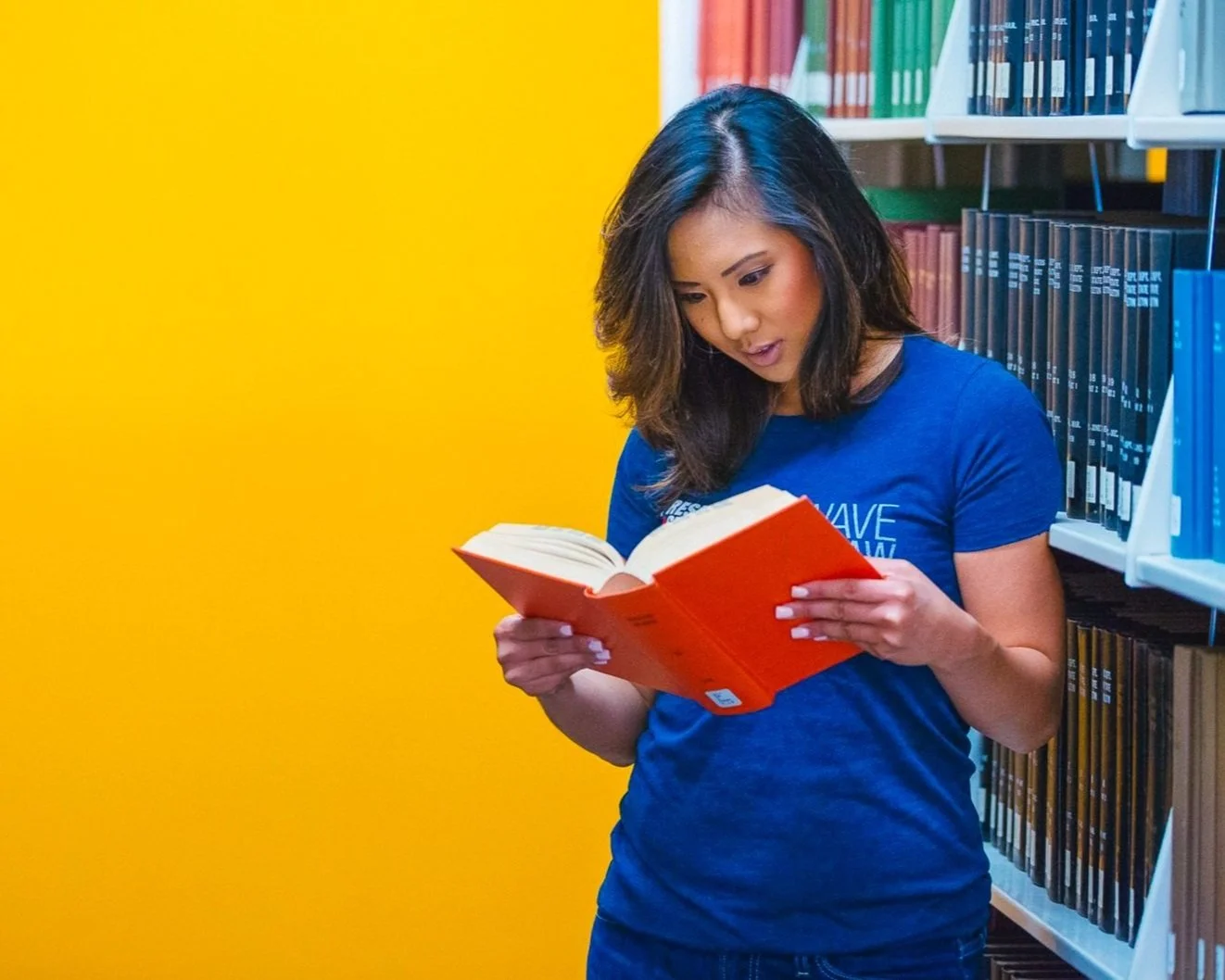 A young woman with dark hair in a blue shirt reading a book in a library with a yellow wall and rows of shelves filled with books.