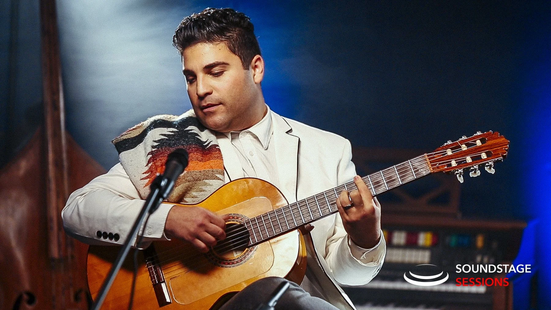 A man in a white suit jacket playing an acoustic guitar in a recording studio with blue lighting. There is a microphone in front of him and a corner of a keyboard is visible in the background. The logo "Soundstage Sessions" is in the bottom right corner.