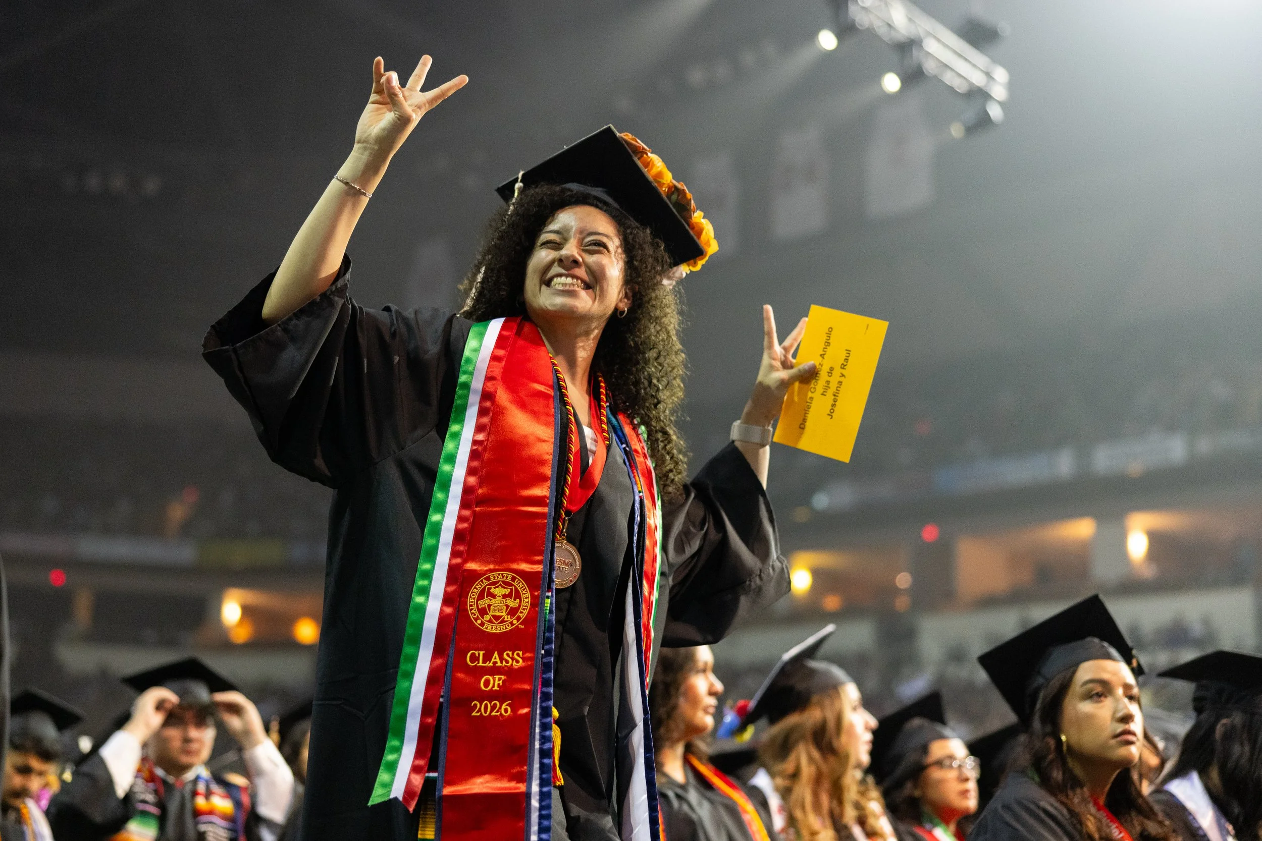 A woman in graduation attire smiling and waving at her graduation ceremony, holding a yellow card, with other graduates seated behind her.