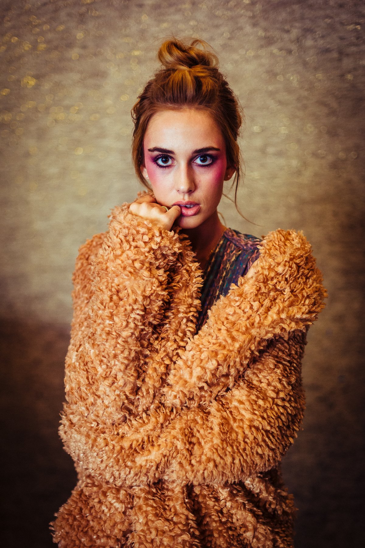 A young woman with fair skin and blue eyes looking directly at the camera. She has pink eye makeup and her hair styled in a messy bun. She is wearing a fluffy, textured tan coat and has her hand near her mouth against a sparkly, silver backdrop.