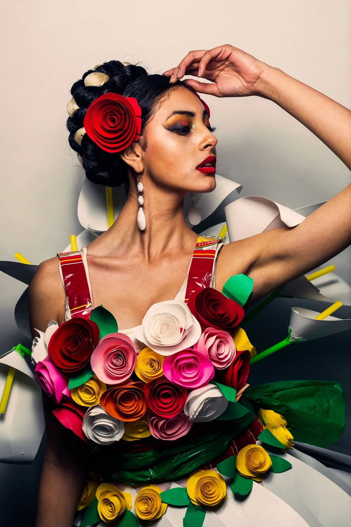 Fashion model wearing a dress decorated with colorful paper roses and flowers, with bold makeup and a red rose hair accessory.