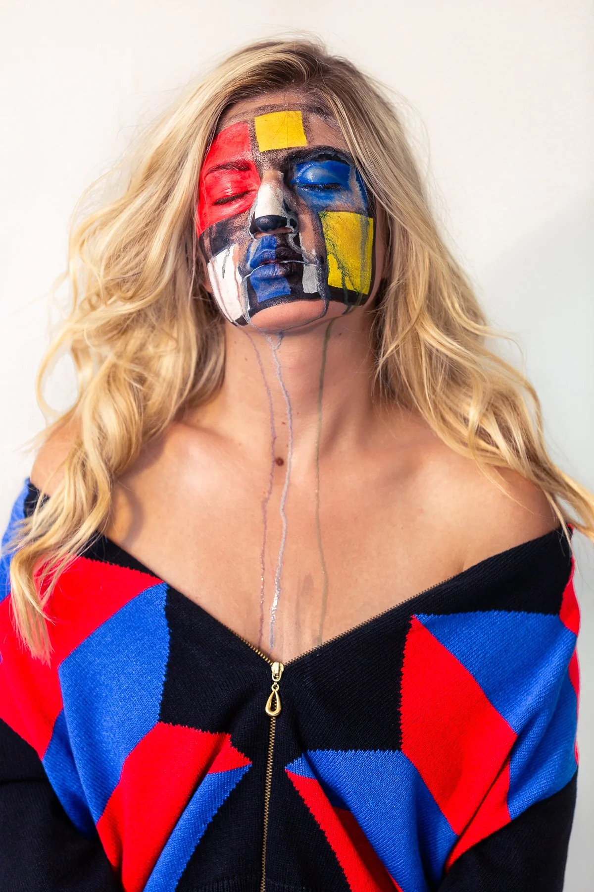 Woman with blonde hair and a black, red, and blue sweater with a zipper, her face painted with abstract, colorful patterns, and paint dripping down her neck.