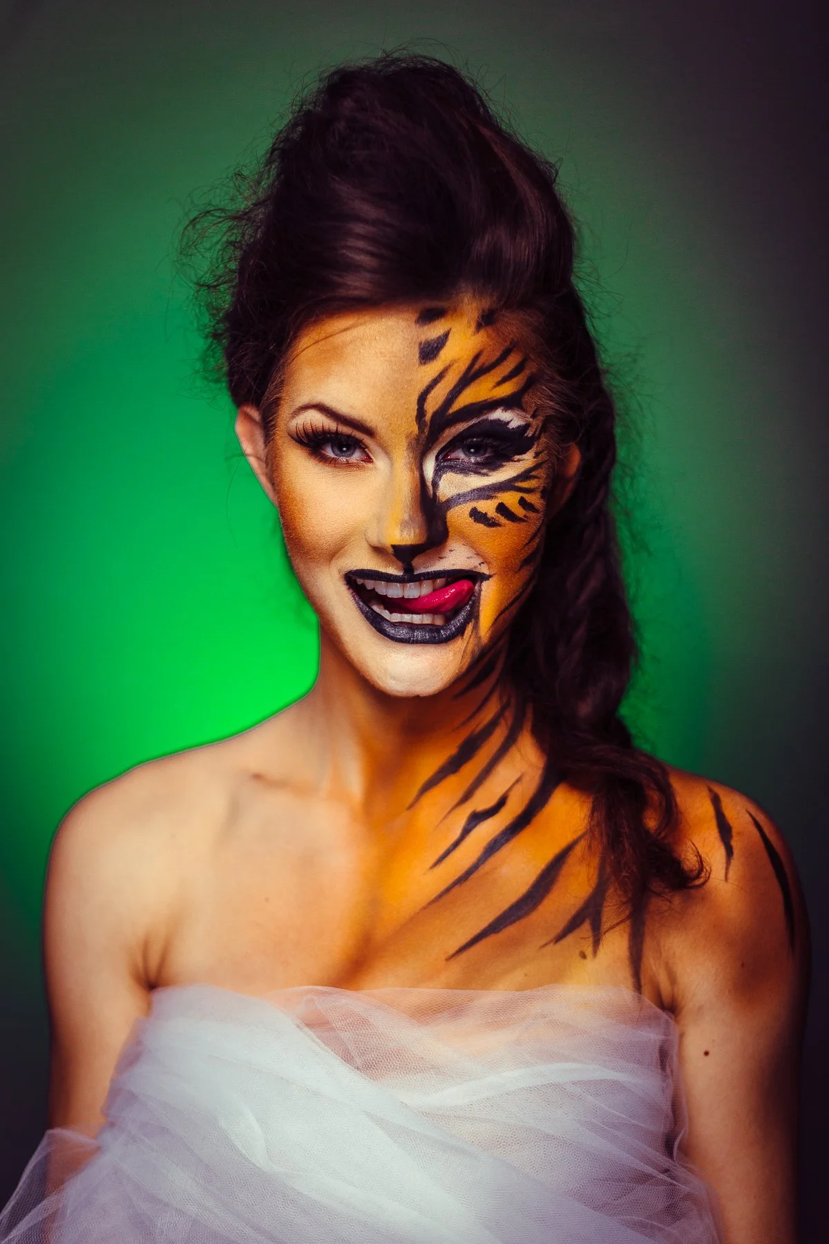 Woman with tiger face paint and tulle wrap, sticking out tongue, against green background.