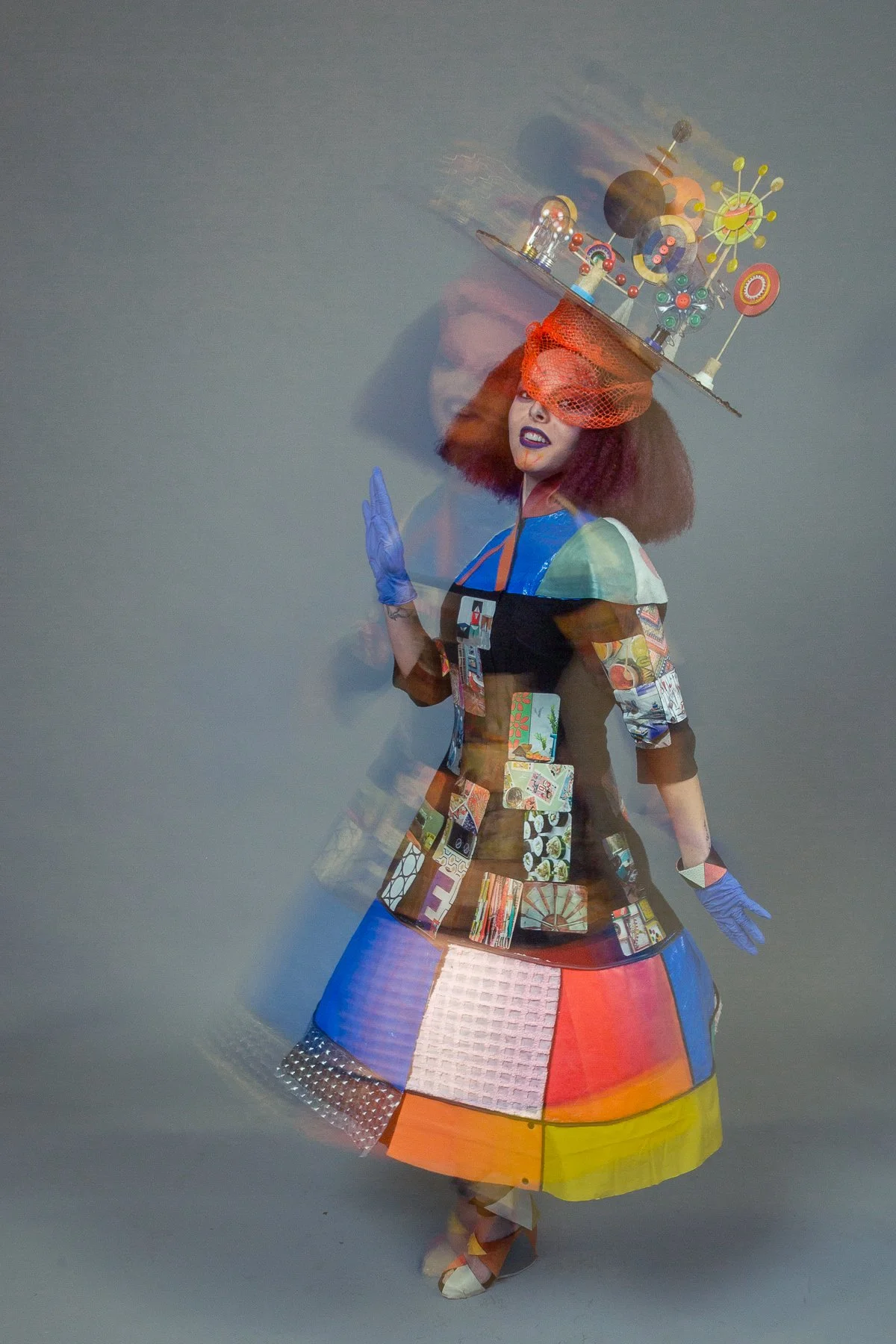 A woman wearing a colorful, patchwork-style dress with a wide skirt, blue gloves, and open-toed shoes. She has vibrant red hair and dark lipstick, with a large, circular hat adorned with various eclectic objects, including small models, toy parts, an