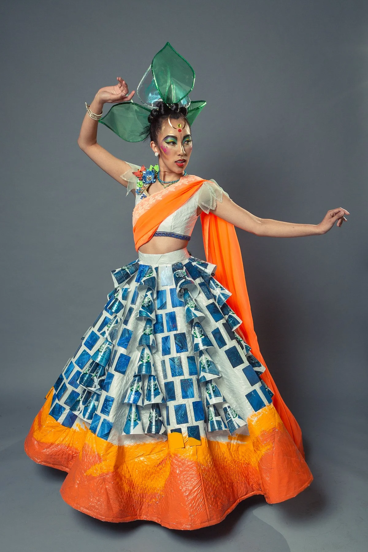 A woman wearing a colorful, elaborate costume with a large green headpiece, orange and white top, and a multi-layered skirt decorated with blue and white patterns, posing against a plain gray background.