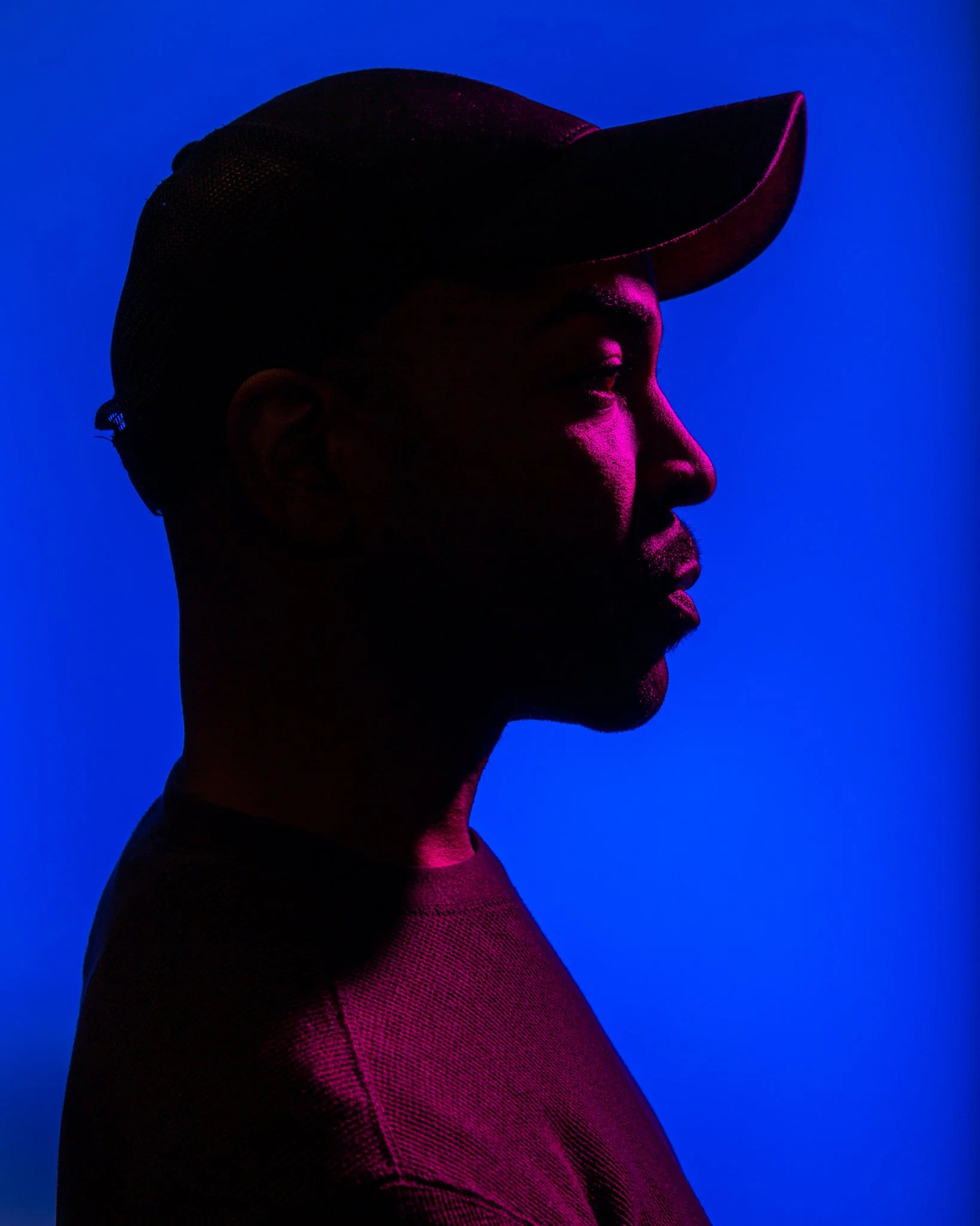 Side profile of a man wearing a baseball cap, illuminated with vibrant purple and pink lighting against a blue background.