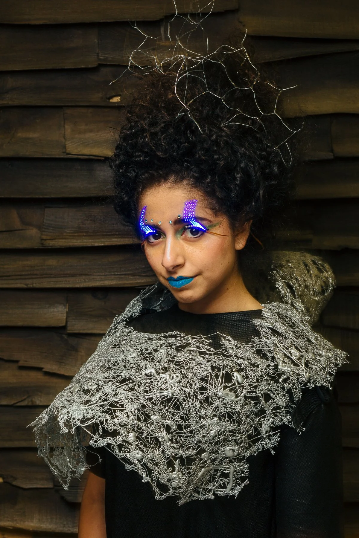 A woman with elaborate digital makeup, bright blue lipstick, and a metallic lace shoulder accessory in front of a dark wooden wall.
