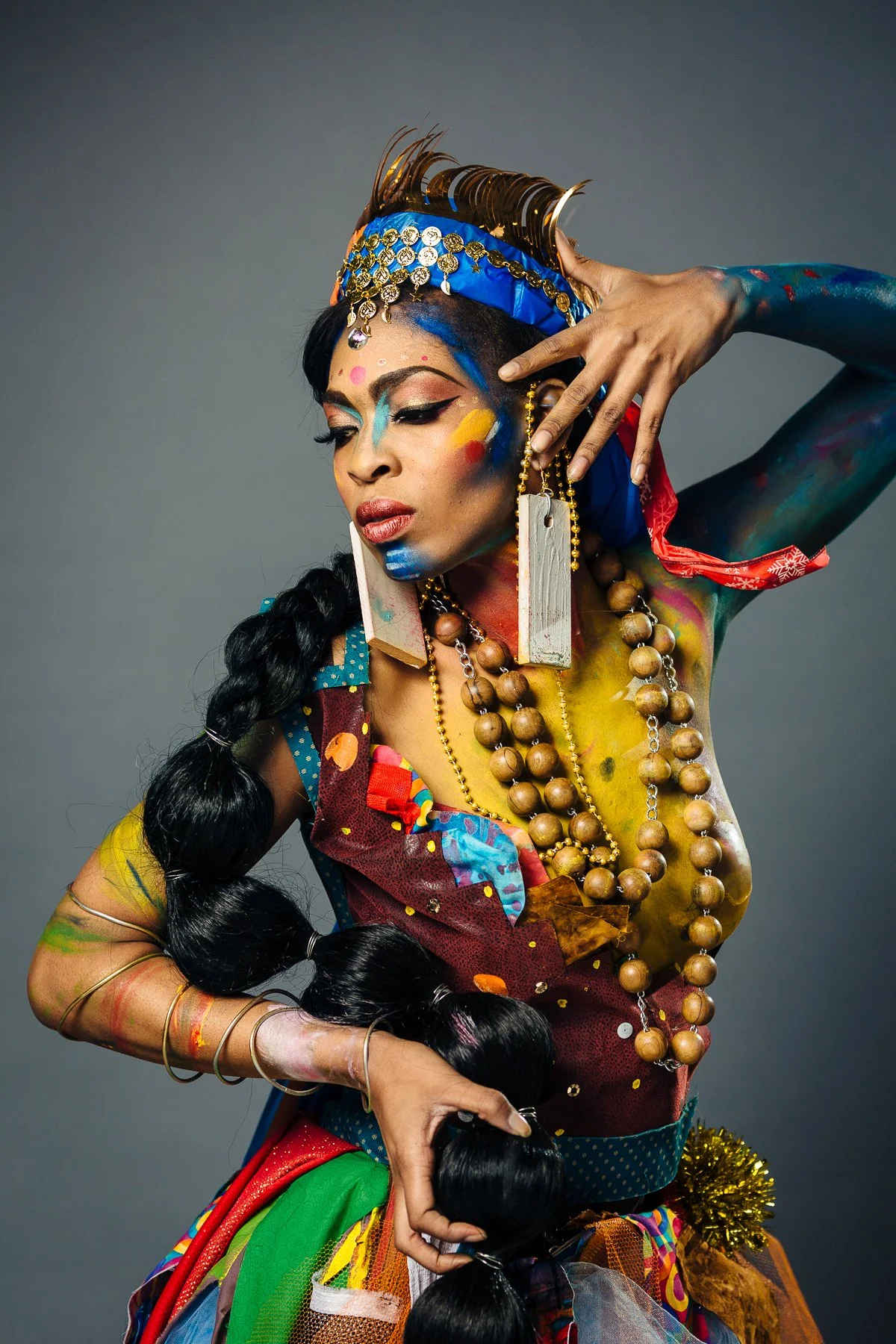 A woman dressed in colorful, elaborate costume with bold makeup and accessories, posing with her hand near her face against a plain background.