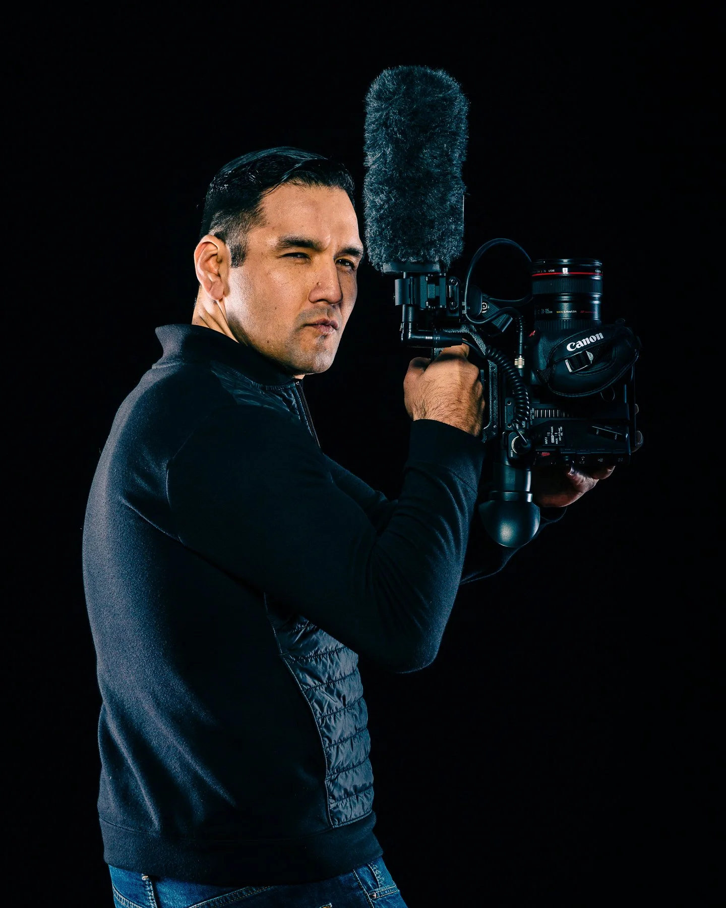 A man holding a professional video camera with a microphone, wearing a black jacket, against a black background.