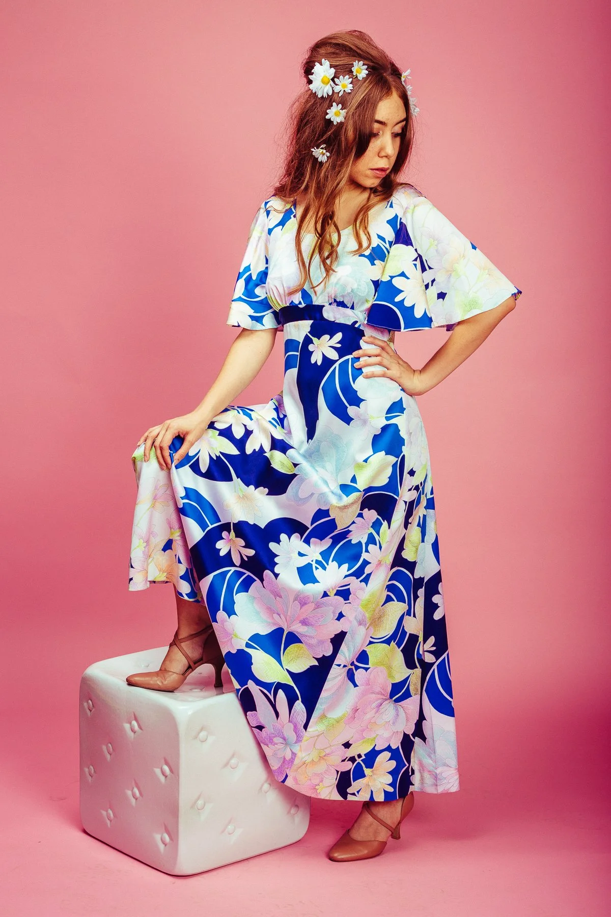 A woman in a floral maxi dress with blue, white, pink, and green patterns, standing on a white upholstered block with a pink background. She has daisy flowers in her hair and is wearing tan high heels.