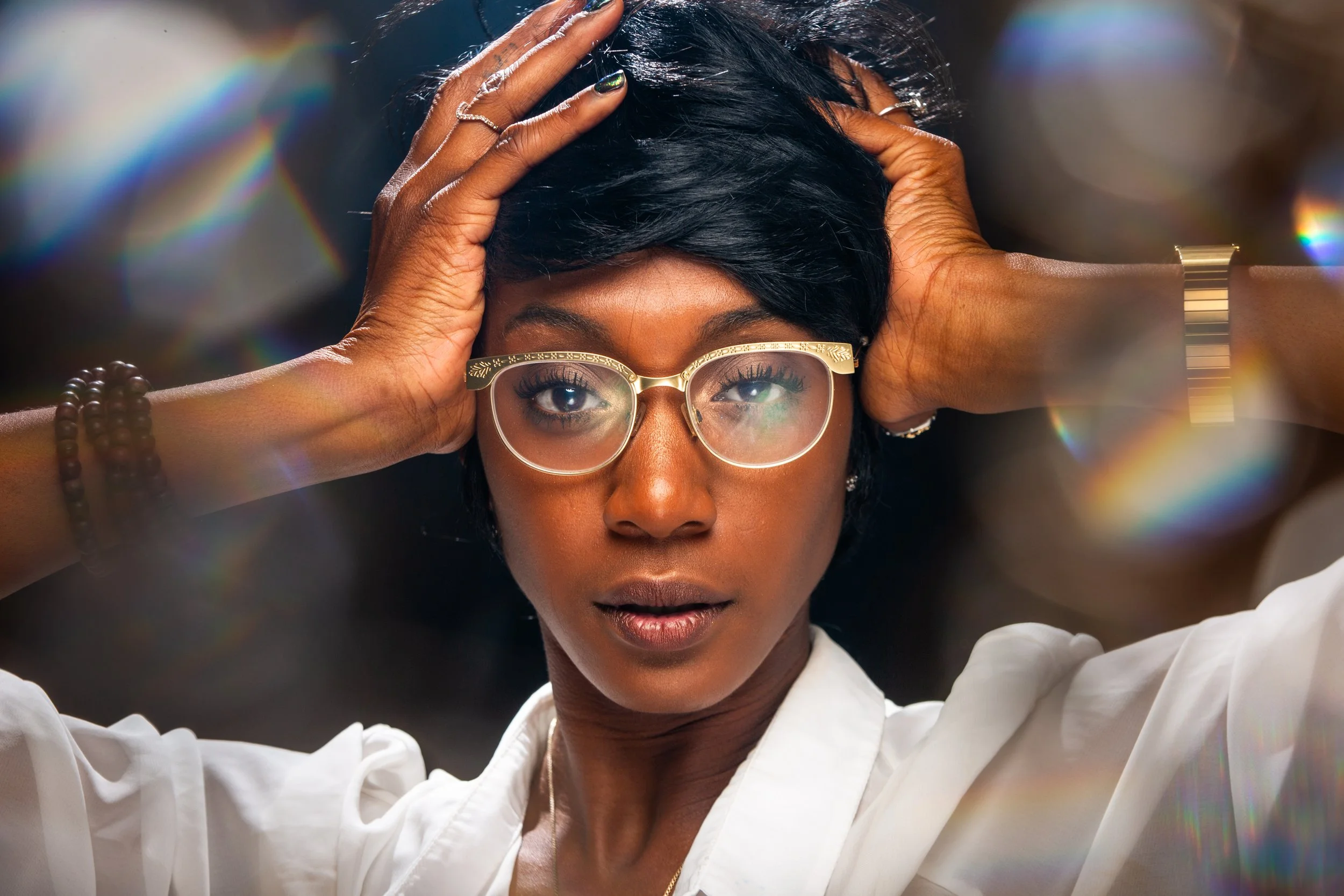A woman with short black hair, wearing glasses, a white shirt, and jewelry, holding her head with both hands, looking directly at the camera with rainbows of light around her.