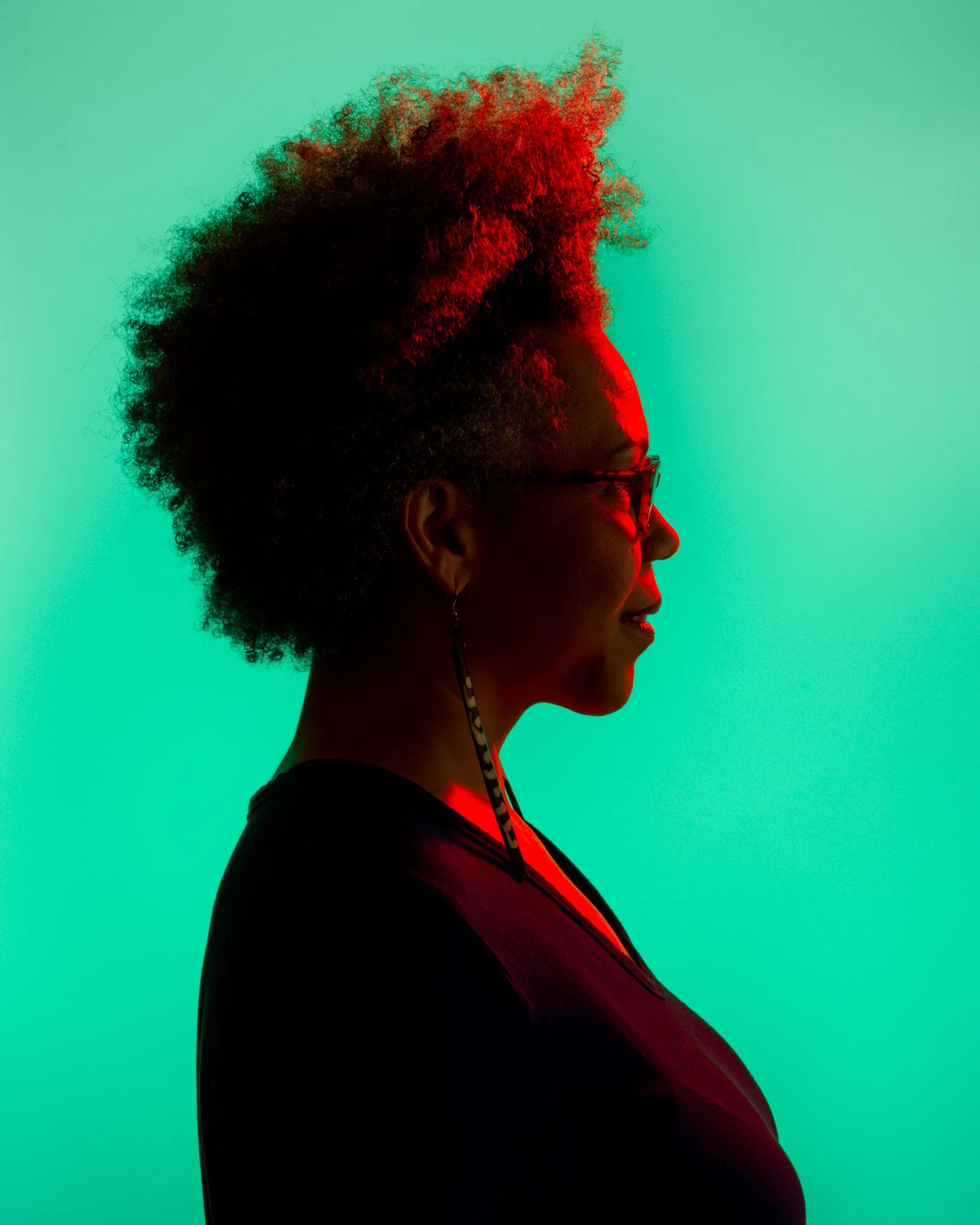 Side profile of a woman with natural curly hair, wearing glasses and a long earring, illuminated with red and teal lighting.