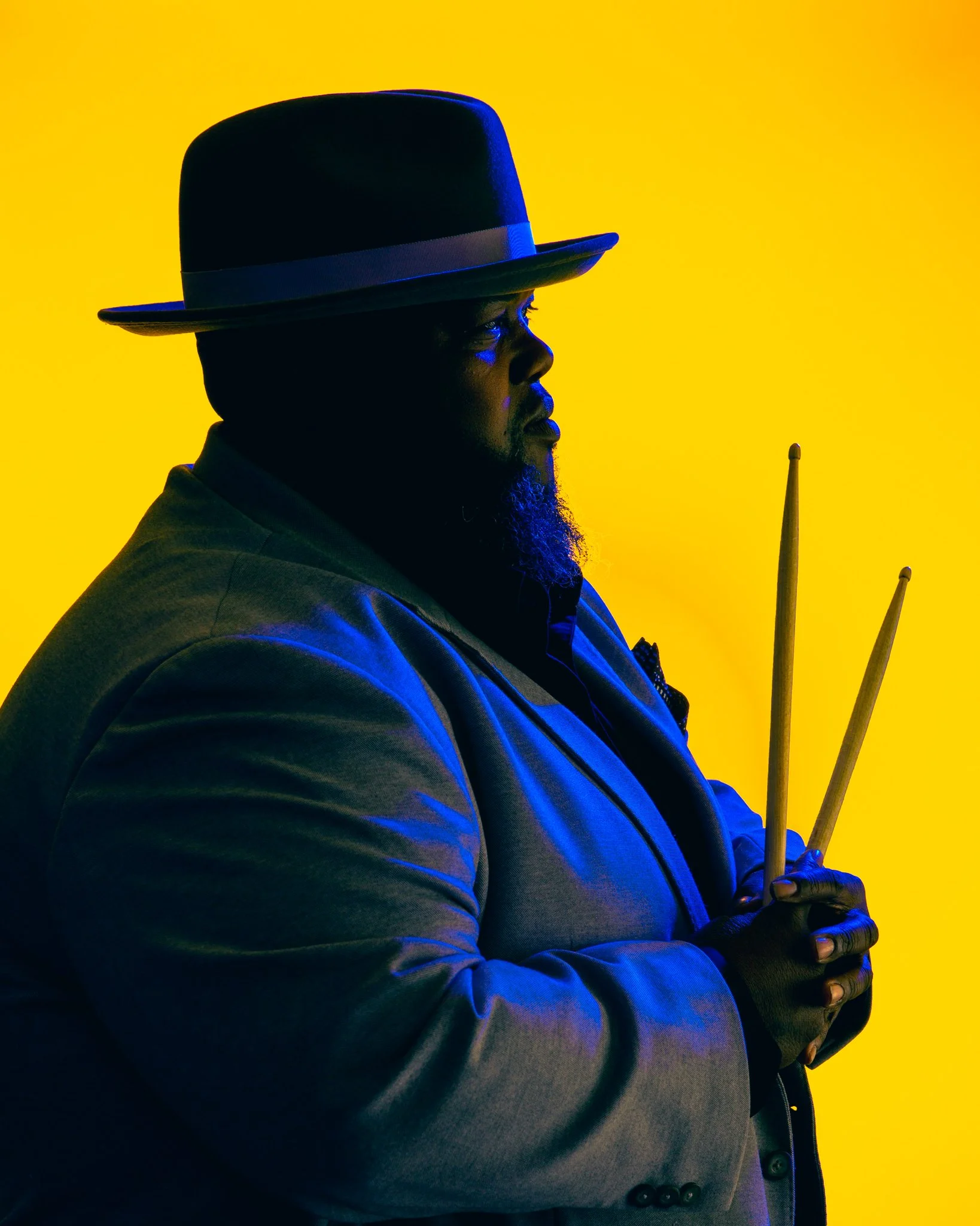Profile of a man wearing a dark fedora hat and a suit, holding drumsticks against a yellow background.