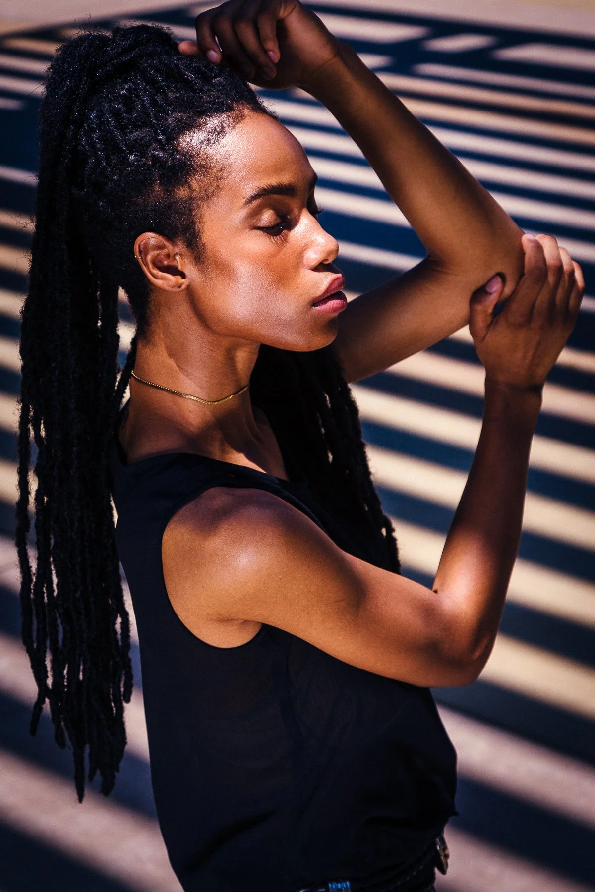 A woman with long dreadlocks in profile, standing outdoors with striped shadows on her skin, her eyes closed, and one arm raised with her hand touching her head.