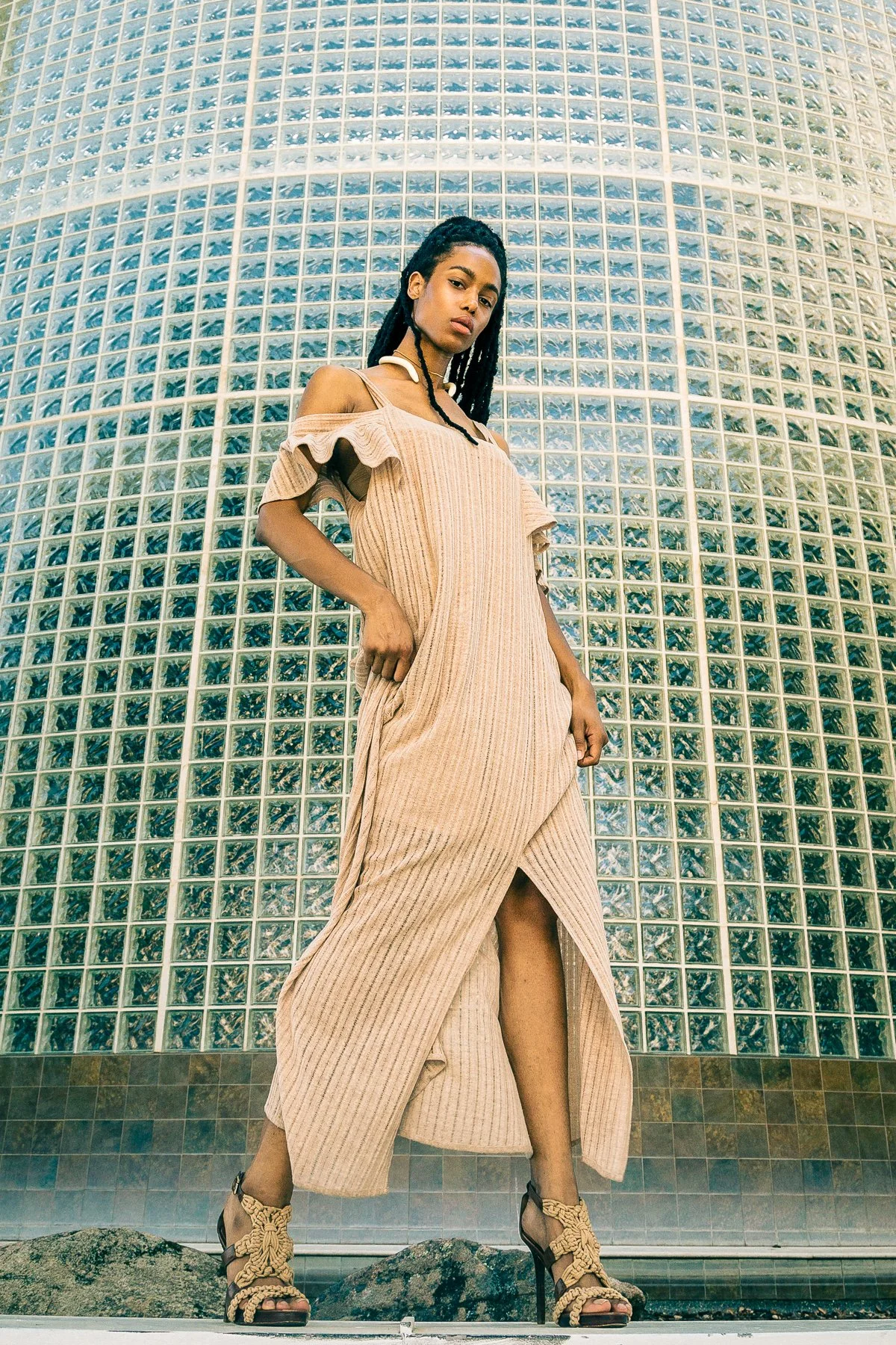 A woman with dark wavy hair styled in dreadlocks posing in front of a large curved glass block wall, wearing a beige off-the-shoulder dress with ruffled sleeves, high heels with knotted straps, and standing on rocks.