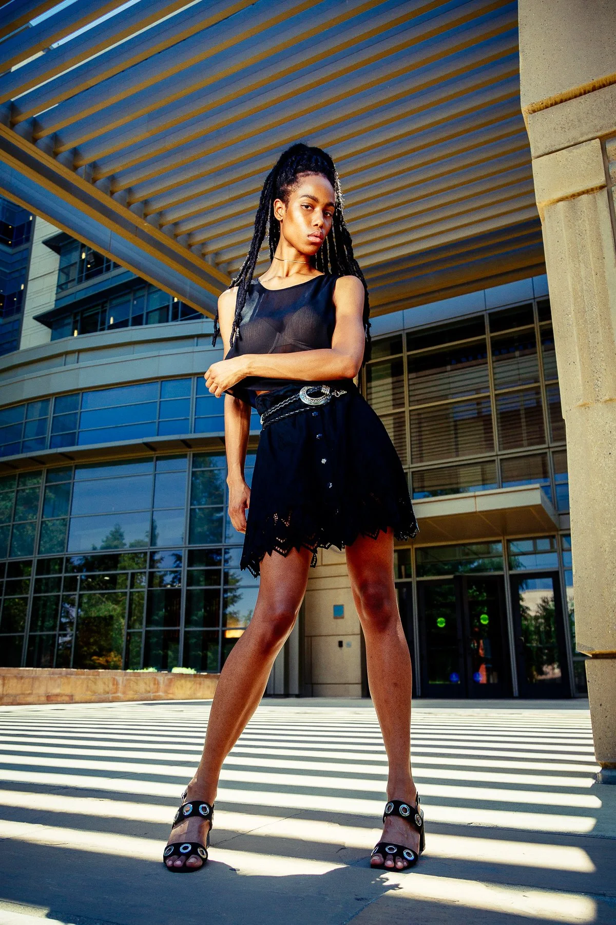 A woman with braided hair standing outdoors in front of a modern glass building, wearing a black sleeveless top, black lace skirt, and high-heeled sandals, striking a confident pose.
