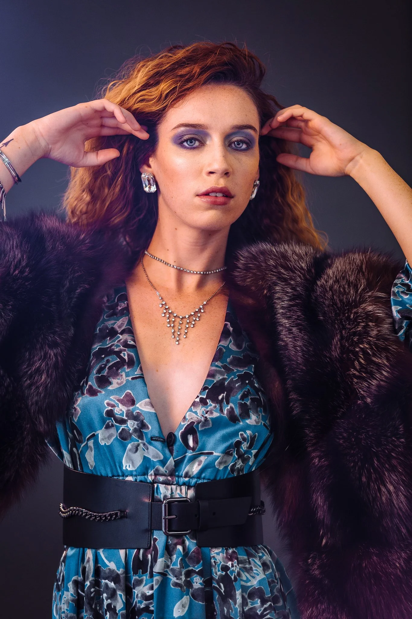 A woman with red wavy hair, wearing a blue floral dress with a black belt, a faux fur stole, and jewelry, posing with her hands near her head against a dark background.