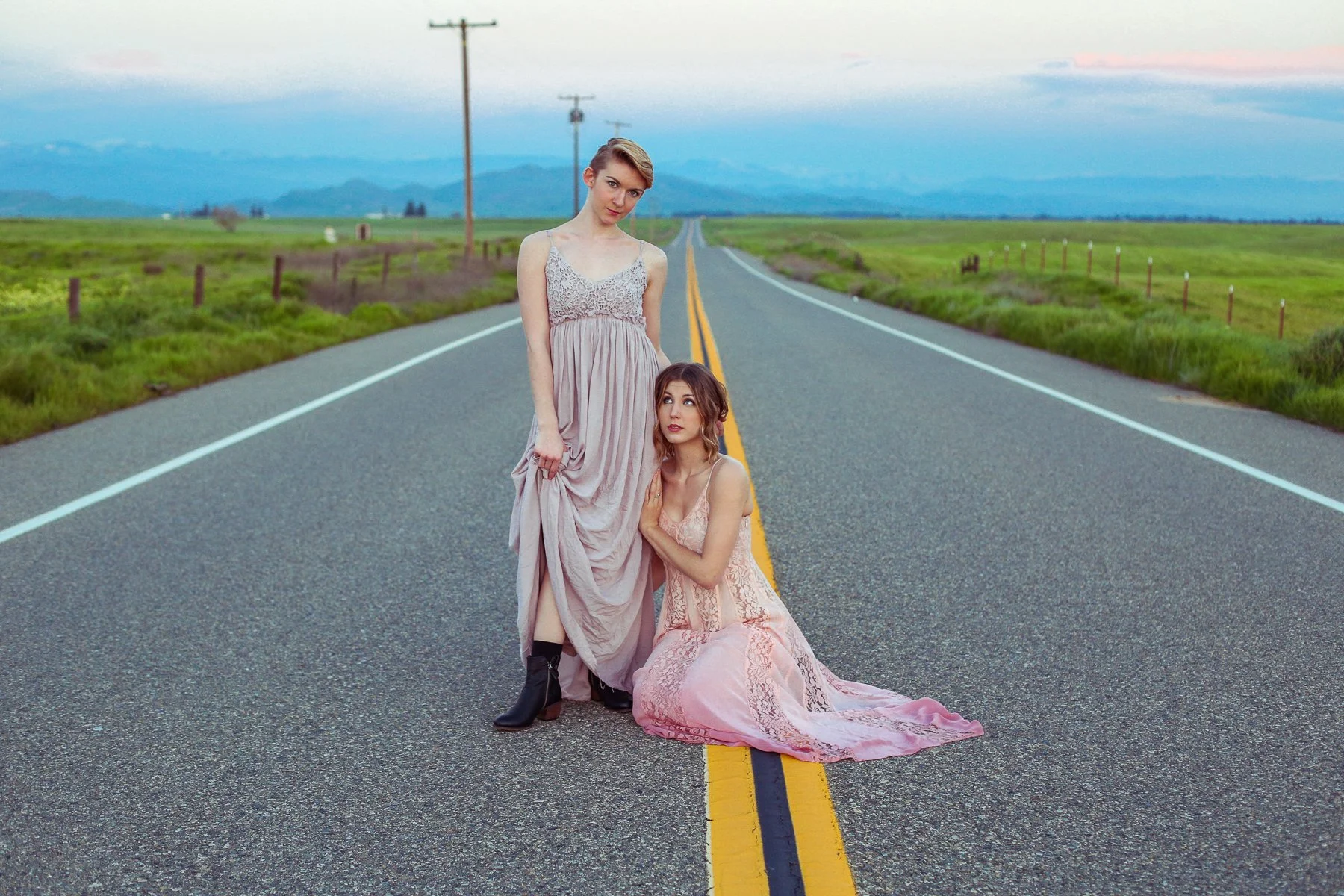 Two women wearing dresses and boots are on an empty road in a rural area with green fields, fences, and mountains in the background during sunset or dusk.