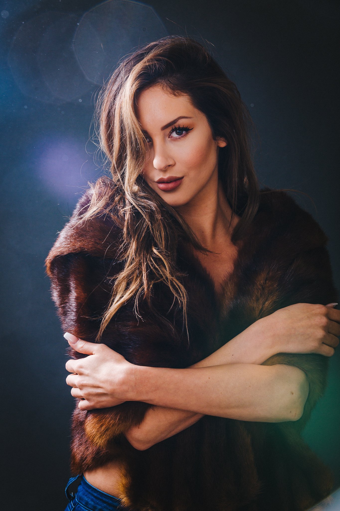A woman with long, wavy brown hair, wearing a fur coat, crosses her arms and looks at the camera against a dark background with lens flare effects.