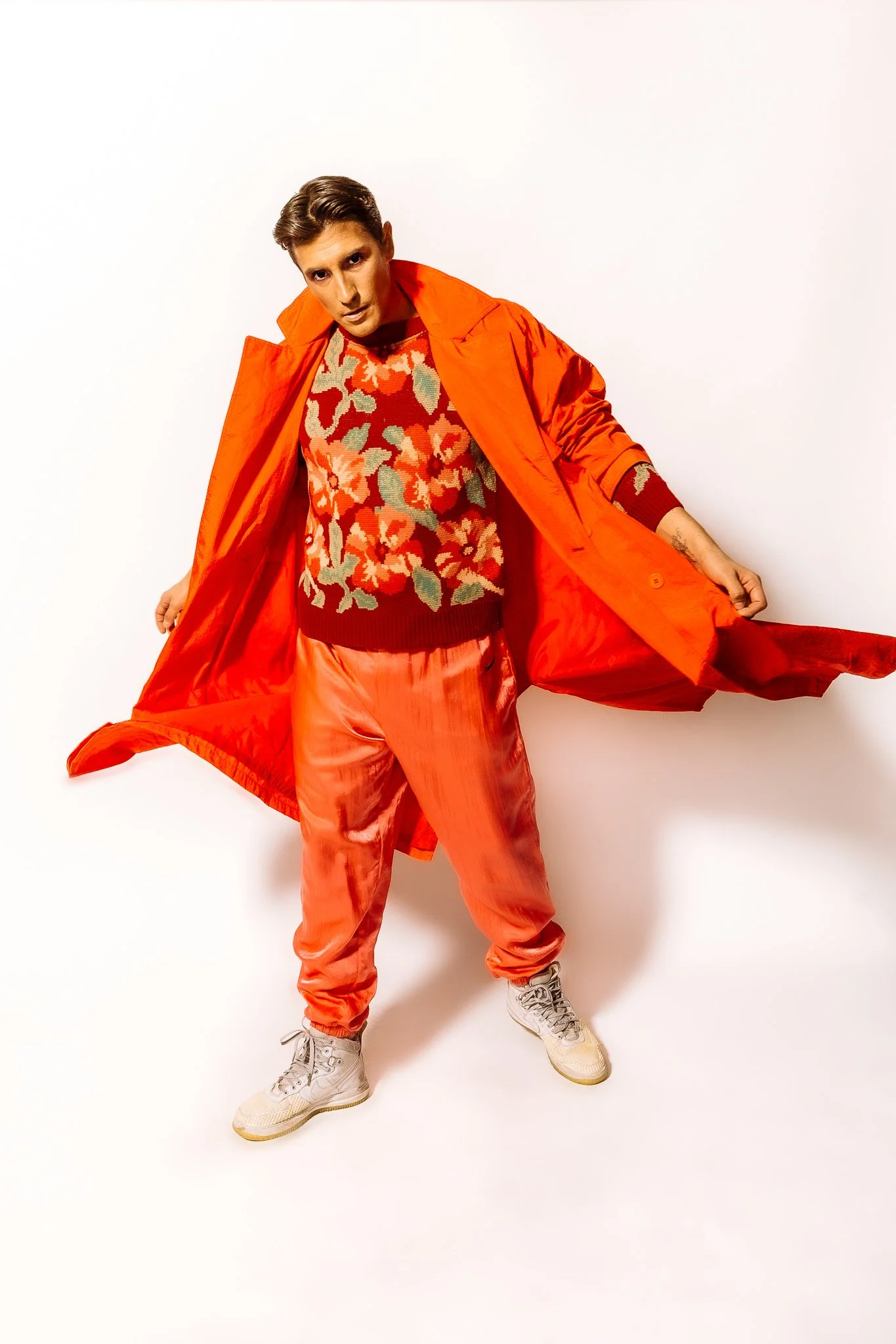A young man wearing an orange coat, floral sweater, and orange pants standing against a white background.