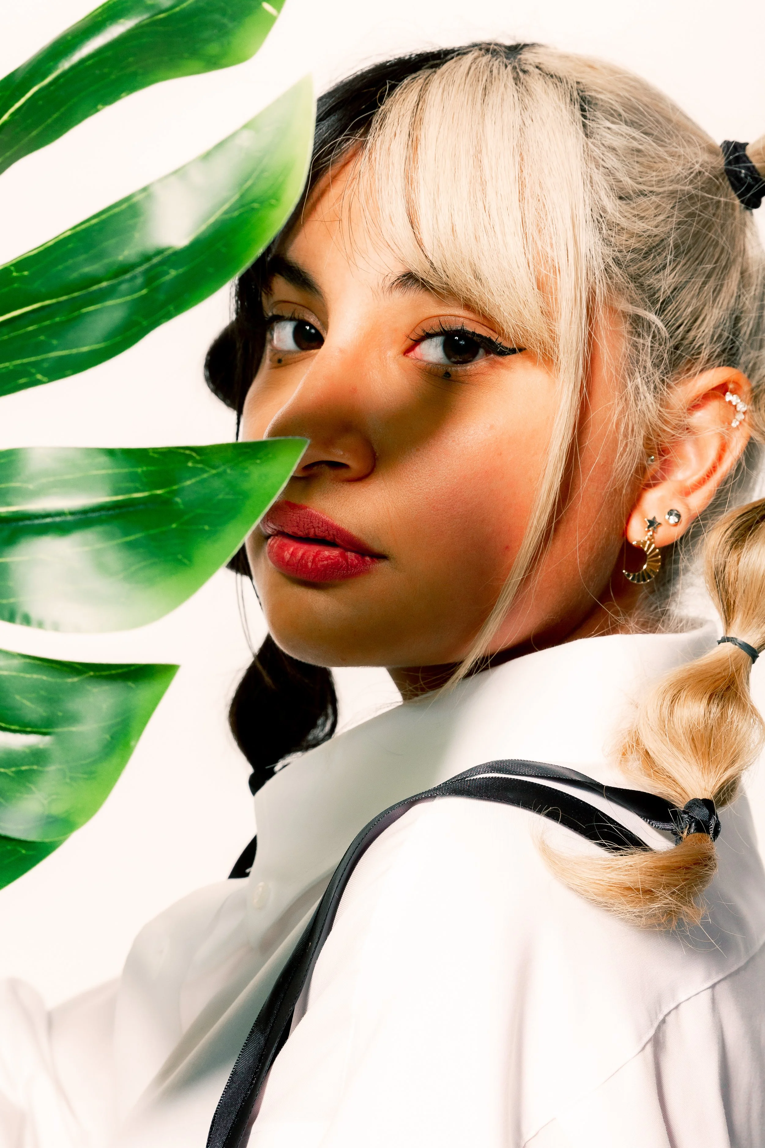 A young woman with blonde and black hair styled in pigtails, wearing multiple earrings, and a white shirt with black accents, posing partially behind large green leaves.