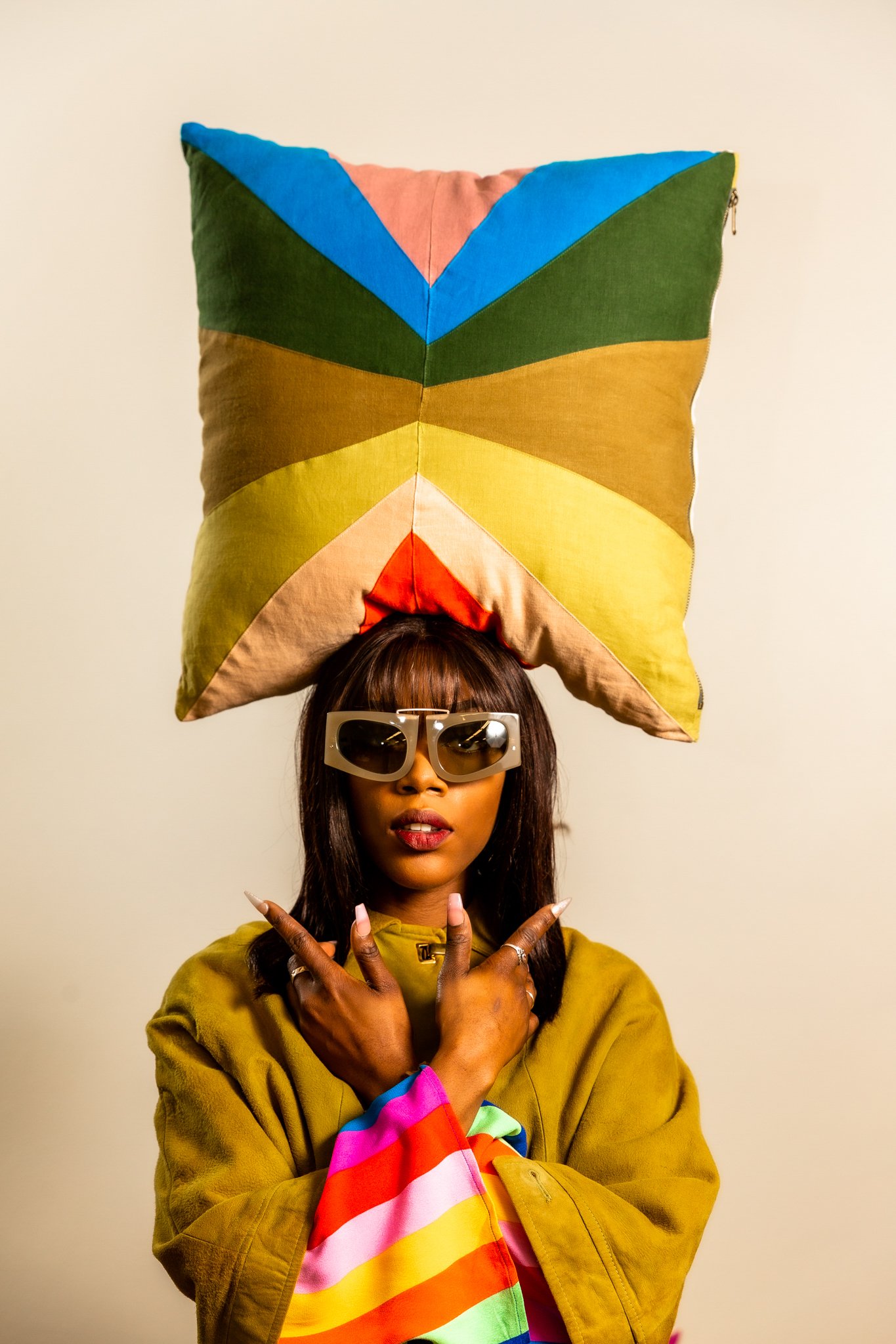 A woman wearing sunglasses, a mustard-colored jacket, and a colorful rainbow-striped shirt. She is balancing a large colorful pillow on her head and making peace signs with her hands.