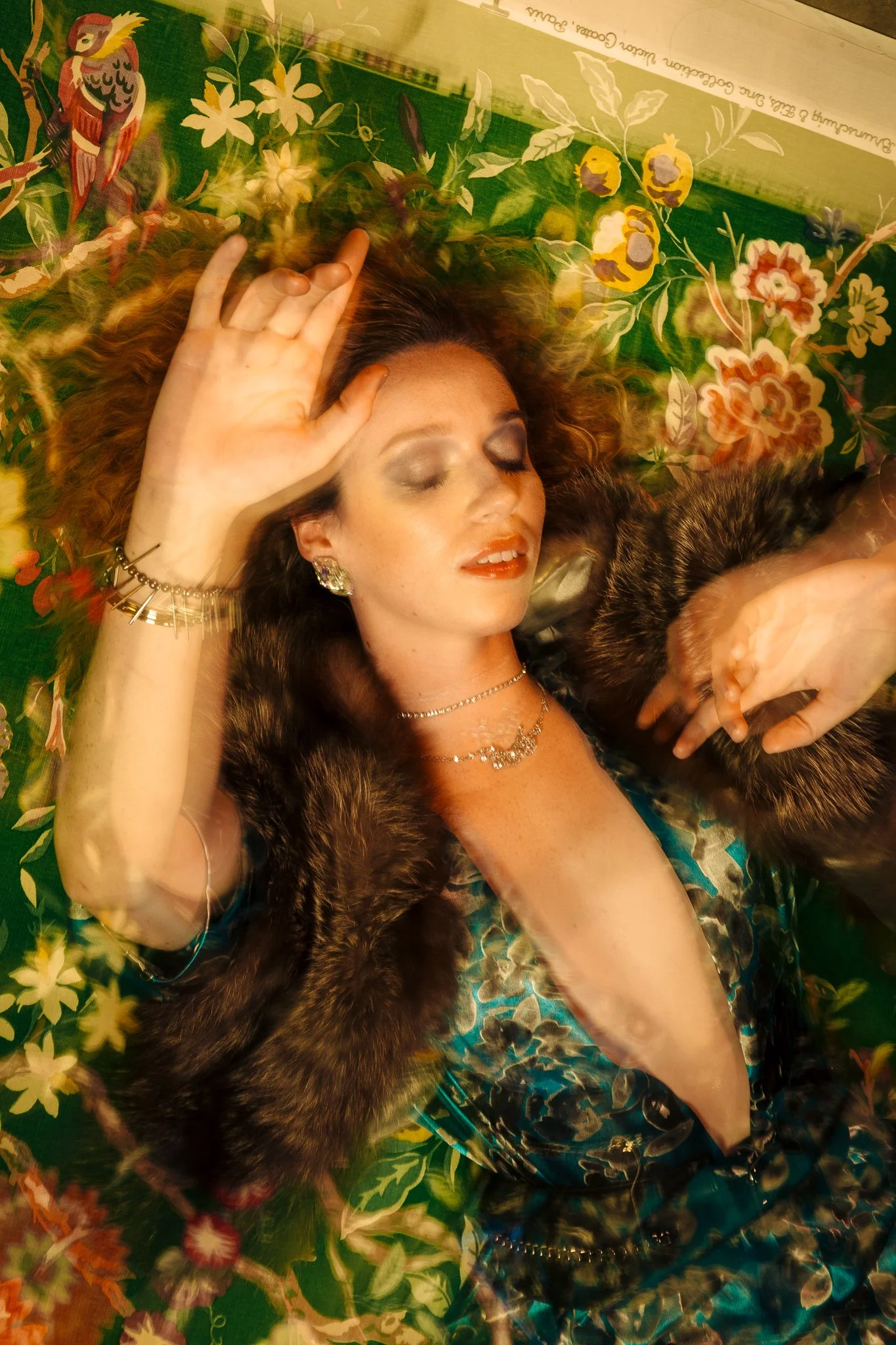 A woman with curly hair lies on a floral-patterned surface with her eyes closed, wearing jewelry, a fur coat, and a silky patterned outfit.