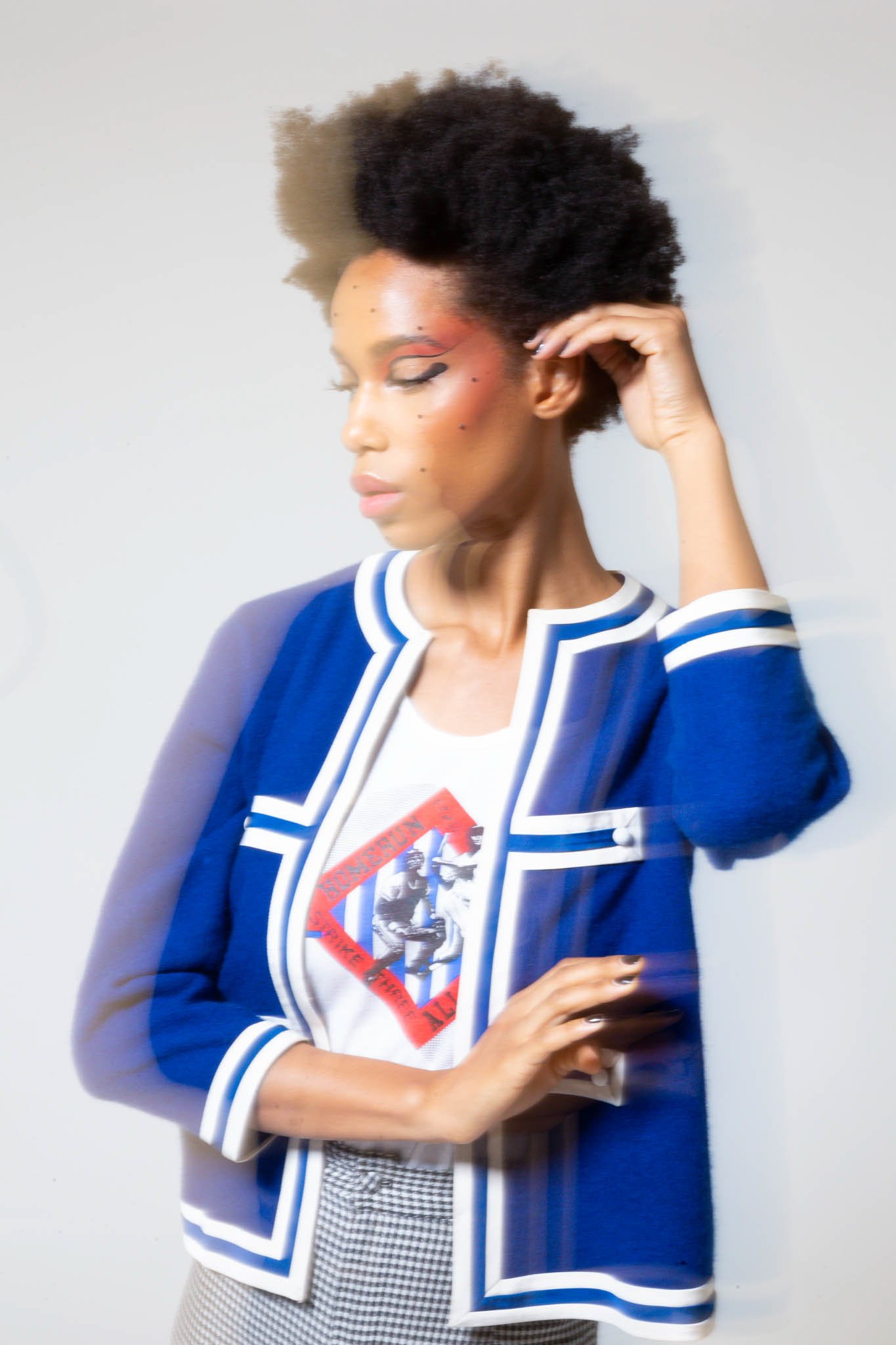 A woman with short curly black hair, wearing a blue blazer with white trim over a graphic t-shirt, and checkered pants, posing with her eyes closed against a plain background.
