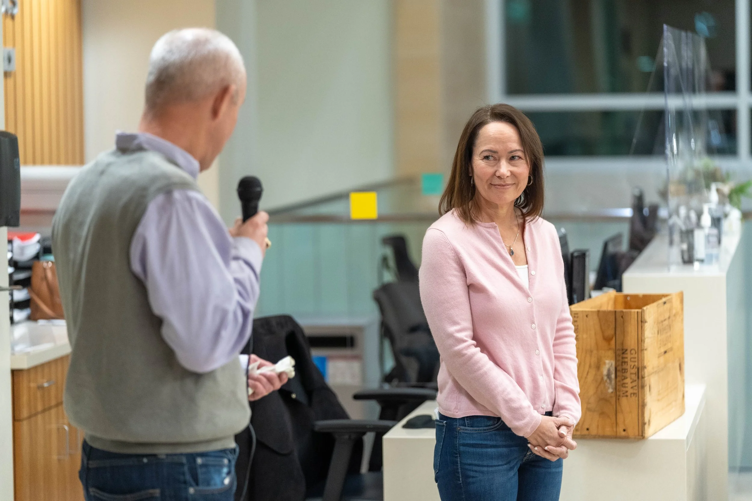 A woman in a pink cardigan and blue jeans looking at an older man holding a microphone during an indoor event.
