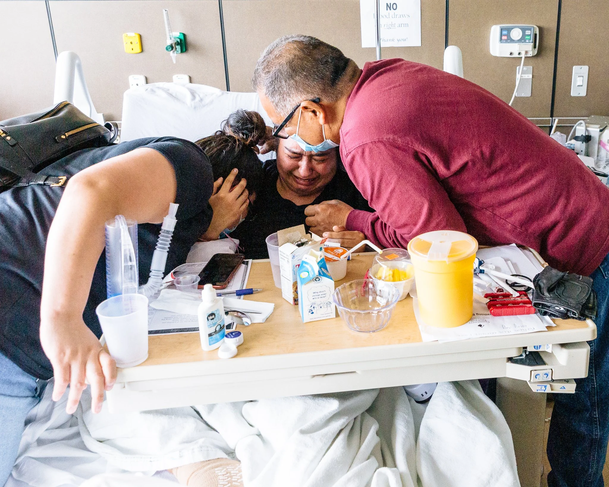 Three people in a hospital room gathered around a patient lying in bed. The patient appears distressed while the two visitors comfort her, with one of them holding her head and the other leaning in close. Medical equipment and supplies are on a table