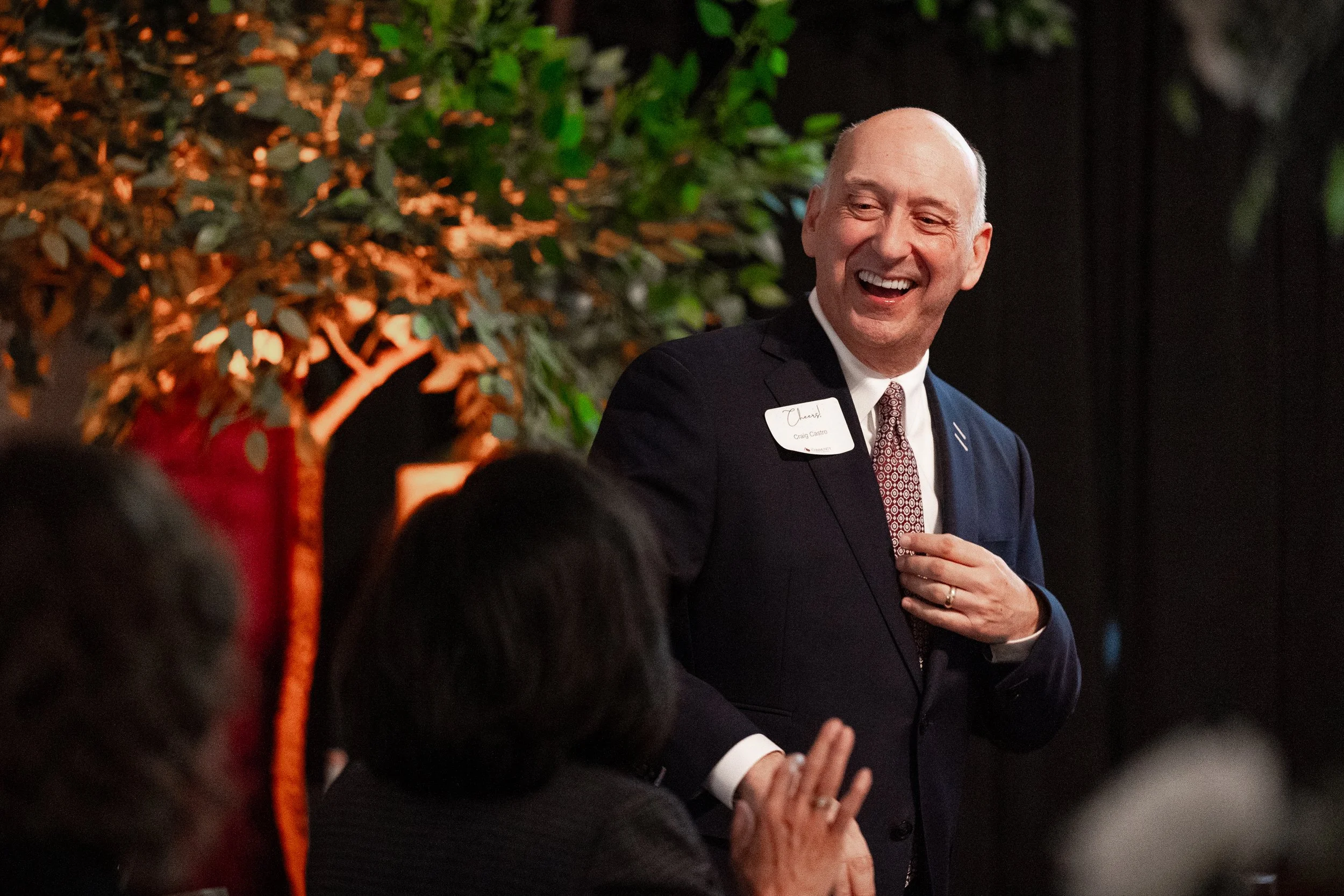 A man in a suit and patterned tie standing and smiling, wearing a name tag, with an indoor background of green foliage and a dark curtain. He appears to be speaking or laughing at an event.