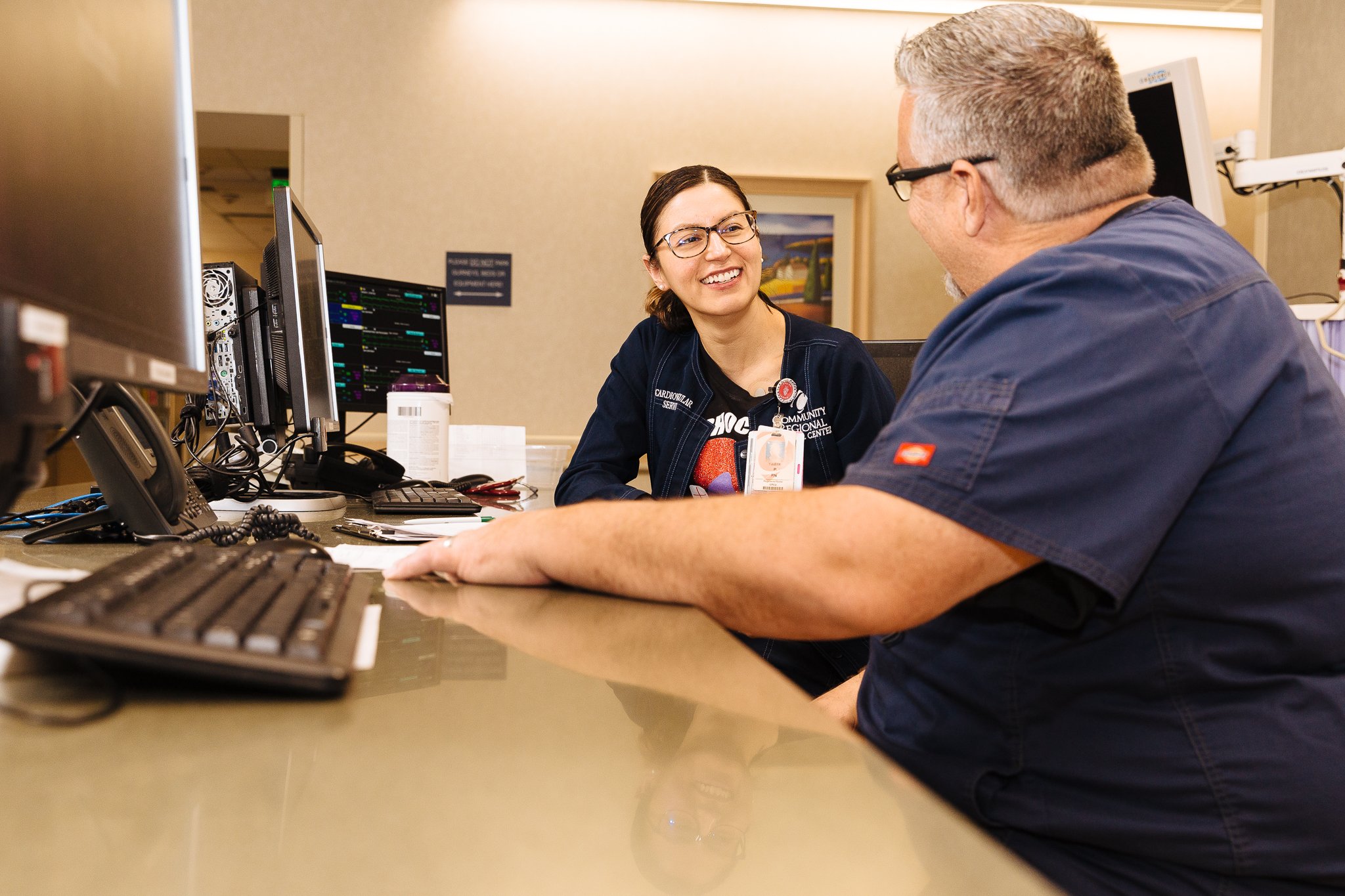 A young woman in medical scrubs and glasses smiling while talking with an older man in a hospital or clinic reception area.