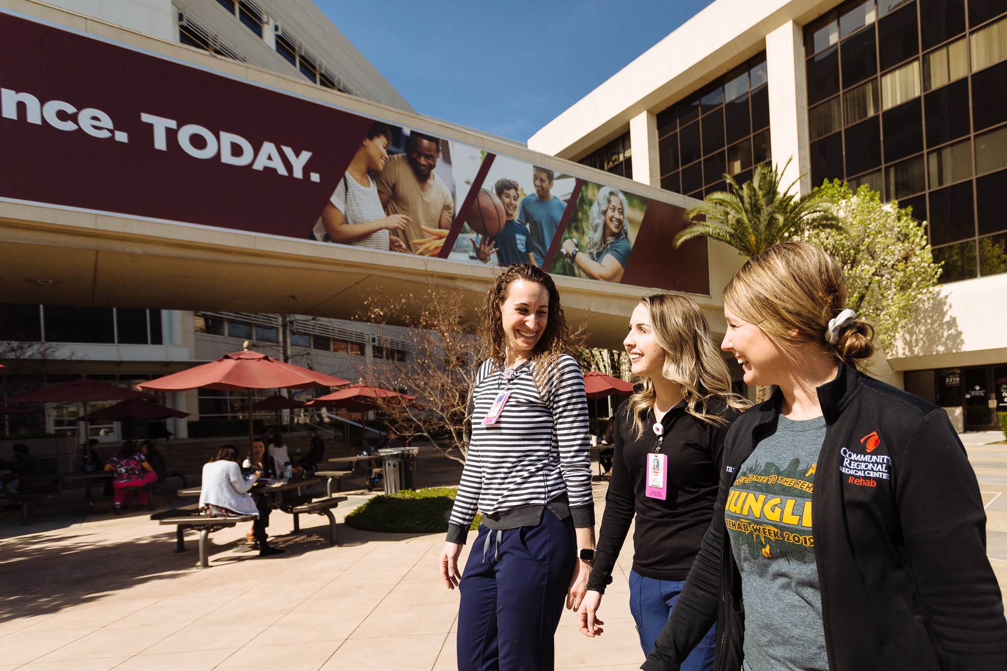 Three women walking and smiling outside a building with a large banner that says 'Today.' in the background. One woman is wearing a striped shirt, another a black jacket, and the third a gray t-shirt with yellow text. Pedestrians are sitting on bench