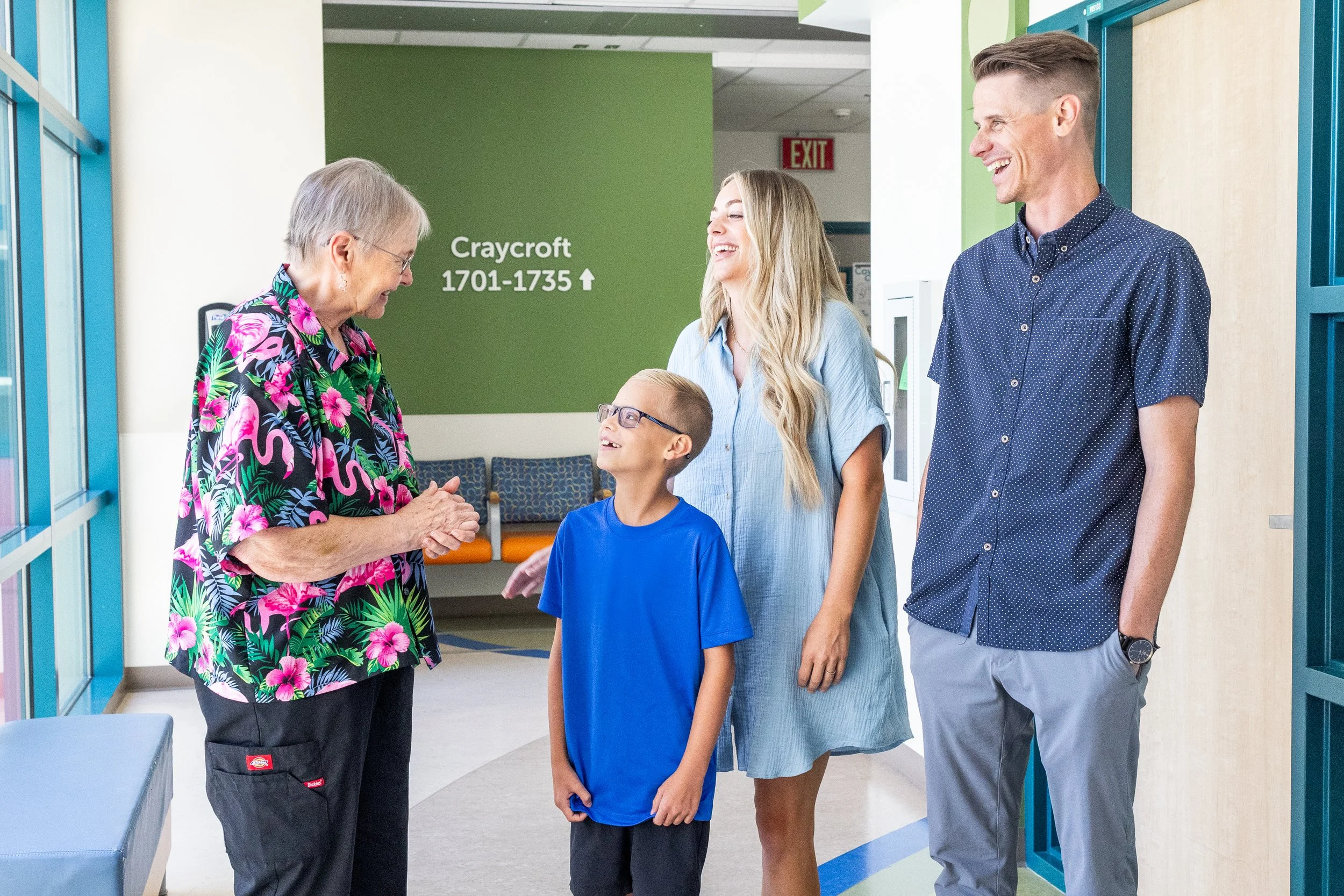 Four people, an elderly woman, a young boy, a young woman, and a young man, are standing and talking in a brightly lit hospital or clinic hallway. The elderly woman is wearing a colorful floral shirt, the boy is in a blue shirt and glasses, the young