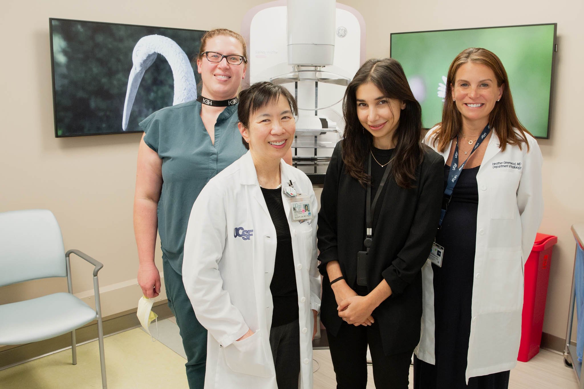 Group of four female medical professionals in a hospital setting, smiling at the camera, with two monitors in the background showing a swan and a green landscape.