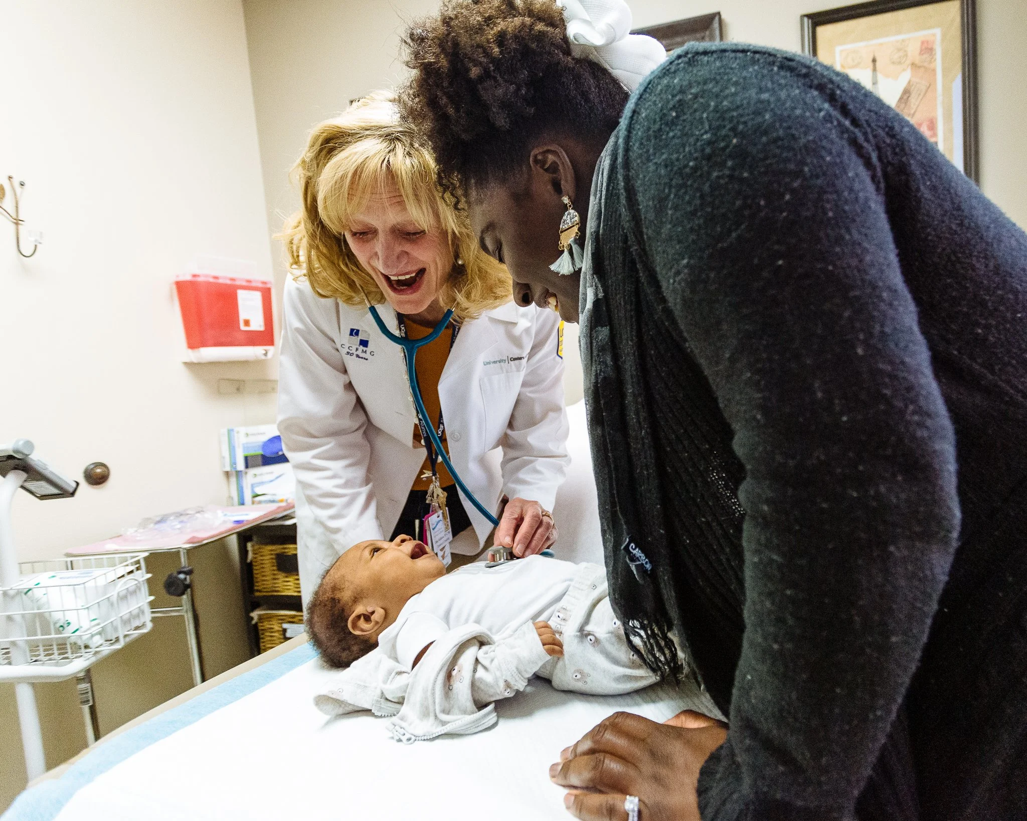 A pediatrician checks a young child's heartbeat with a stethoscope while a woman, possibly the child's mother, looks on with concern in a hospital room.