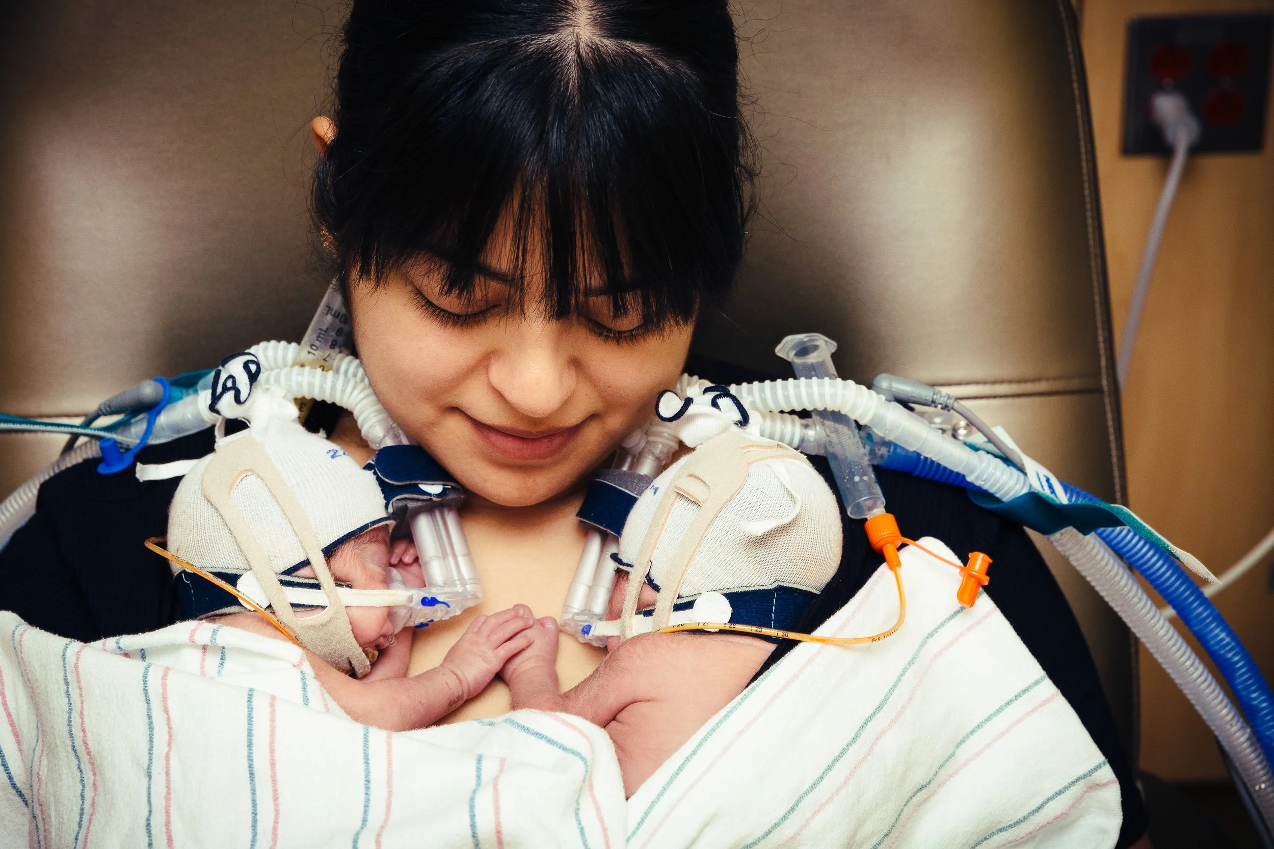 A woman in a hospital bed with a newborn baby and a preemie wearing a cap and breathing tubes, smiling gently.
