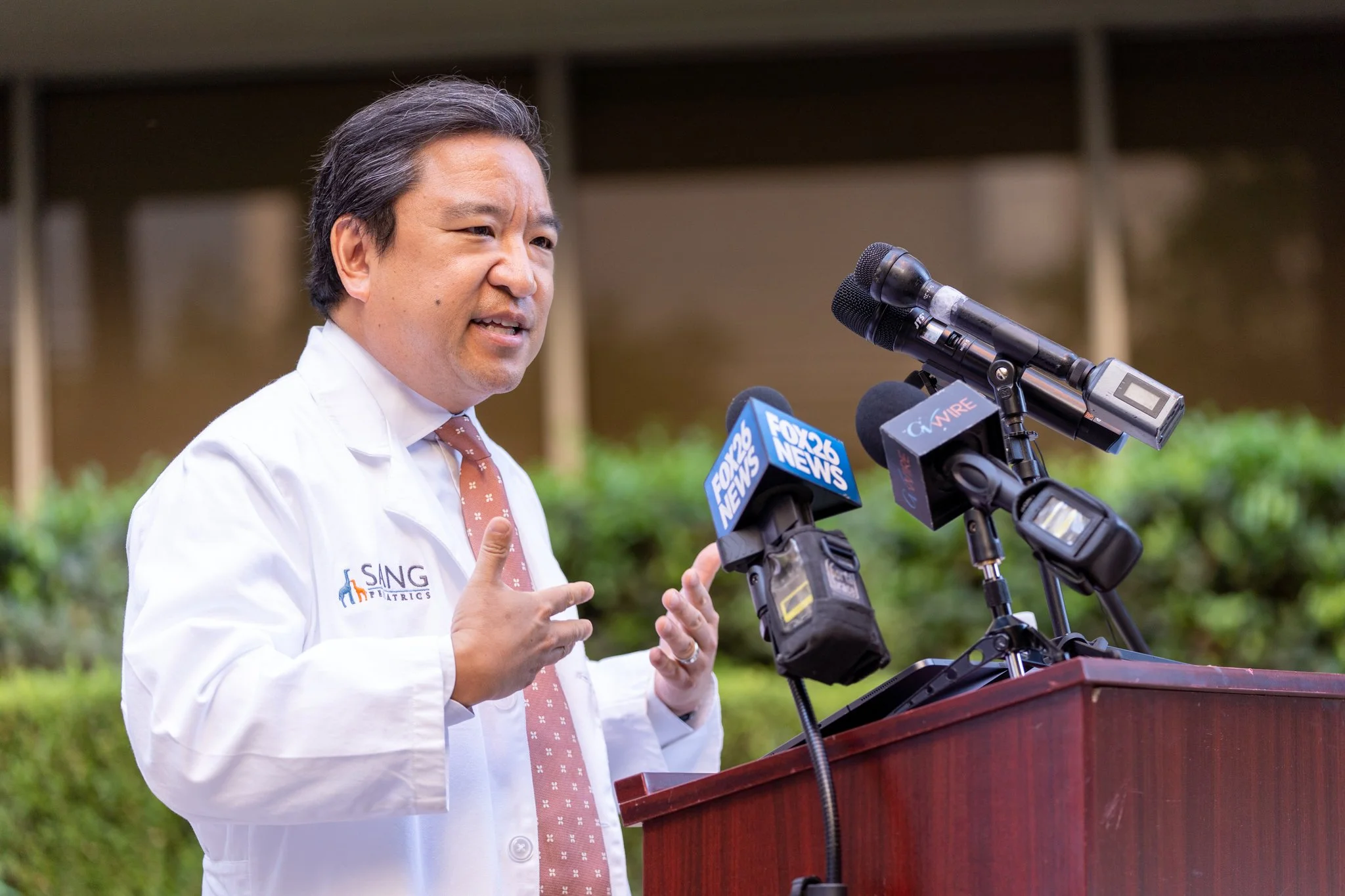 Man giving a speech at a podium with microphones from FOX 26 NEWS and other media outlets, wearing a white lab coat and a pink tie, outdoors with greenery in the background.