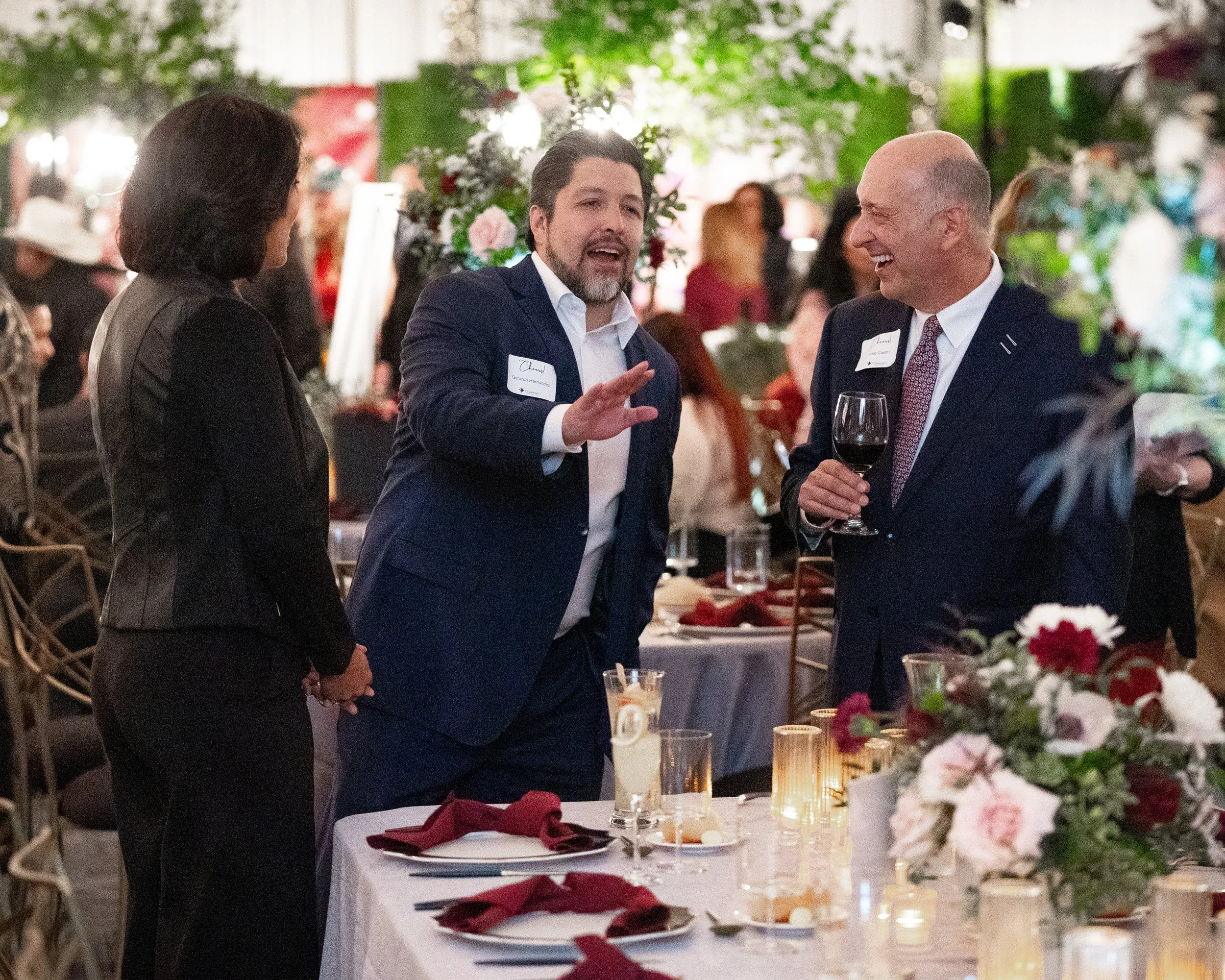 Three people in formal attire at a social event, one man gesturing and another holding a glass of red wine, with floral centerpieces and candlelit tables in the background.