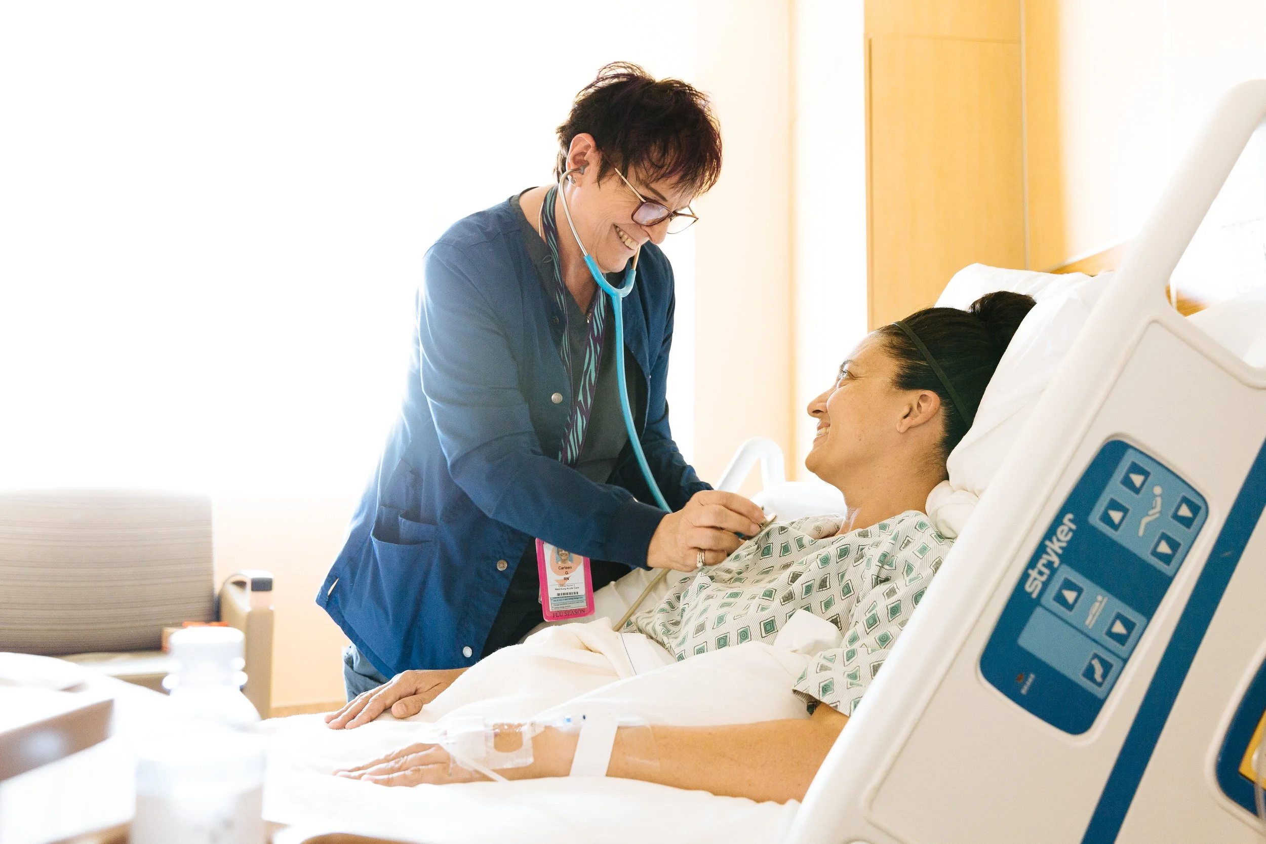 A nurse checks on a smiling female patient lying in a hospital bed.