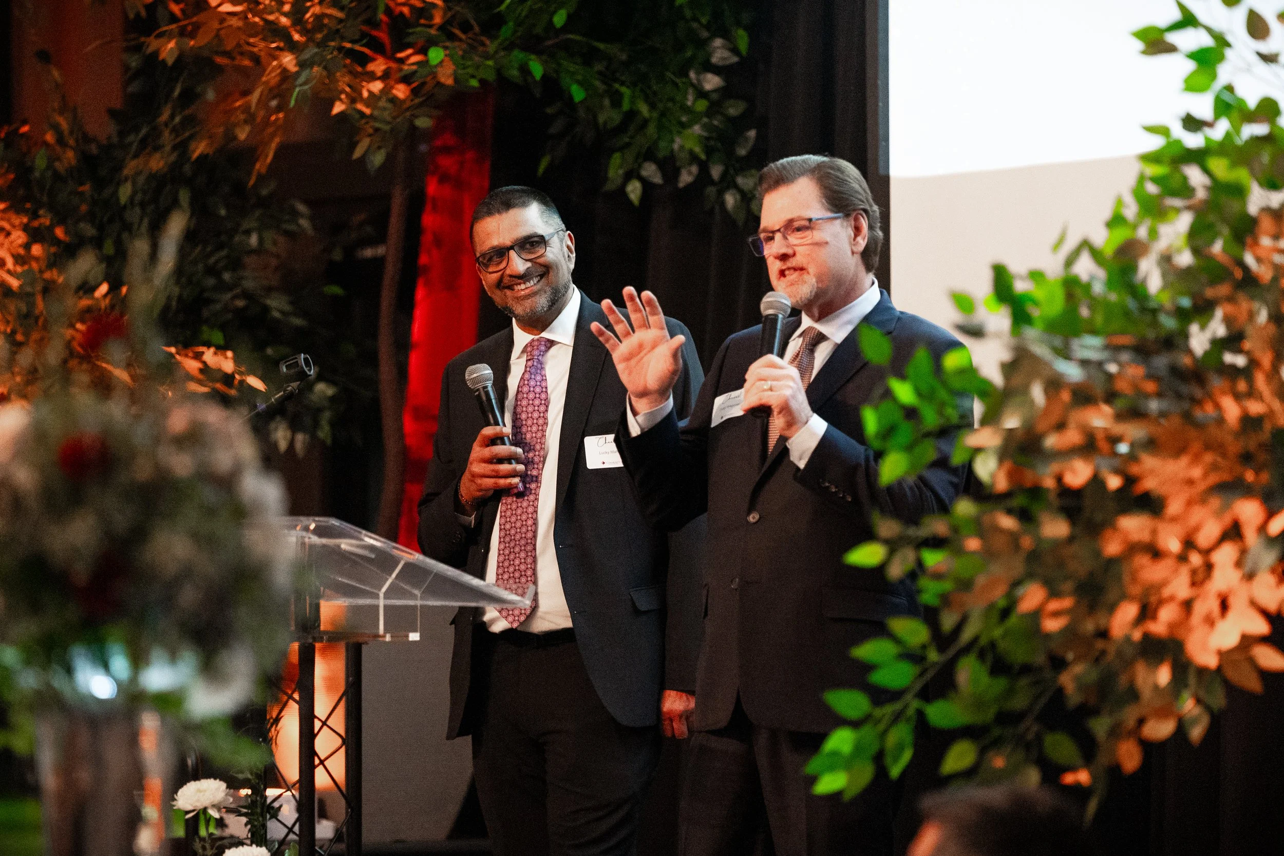 Two men in suits holding microphones, speaking at a formal event with floral decorations and a large screen in the background.