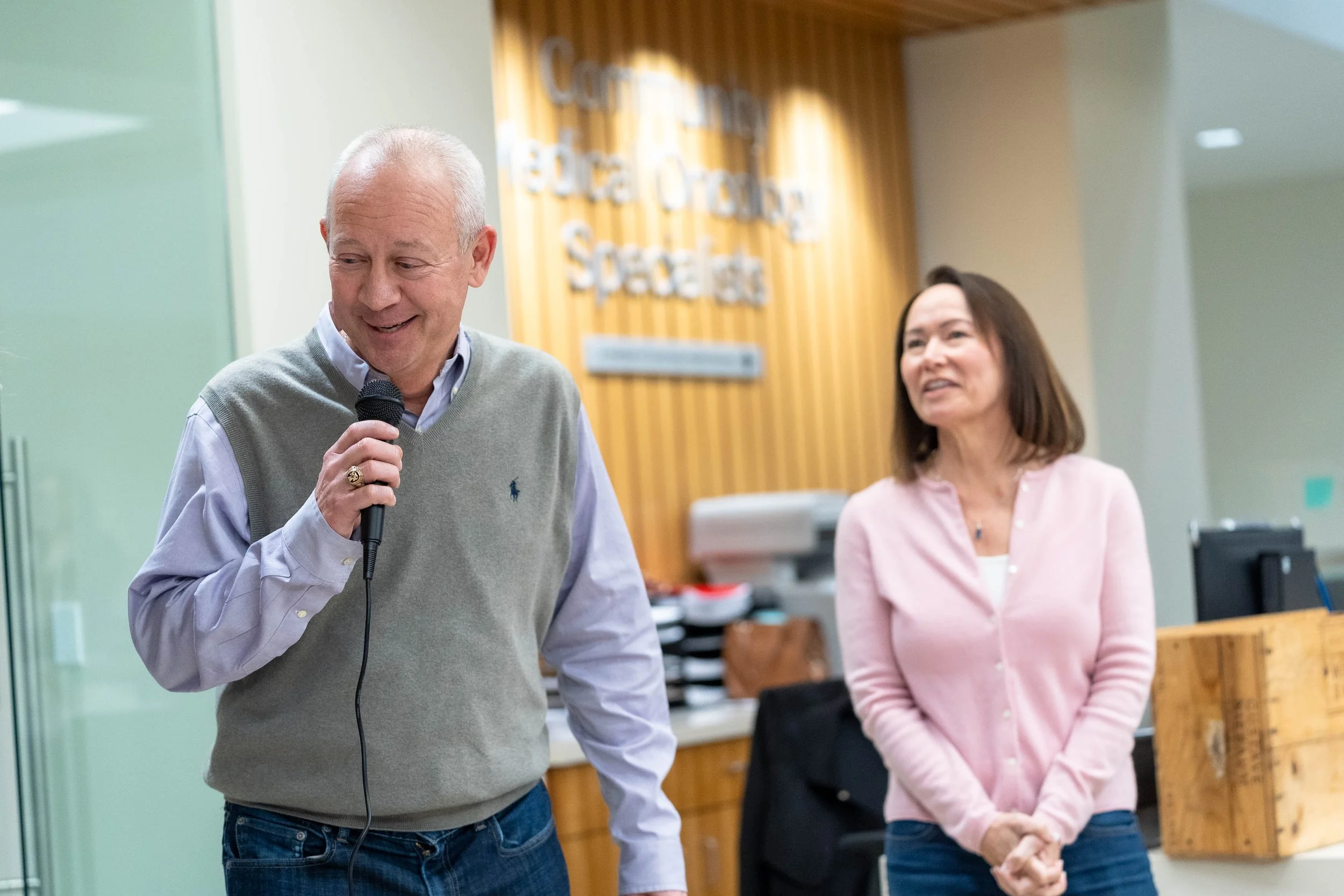 An older man with white hair speaking into a microphone, standing beside a woman with shoulder-length dark hair, in an office setting with a wooden wall and a sign that reads 'Counseling Before School' in the background.