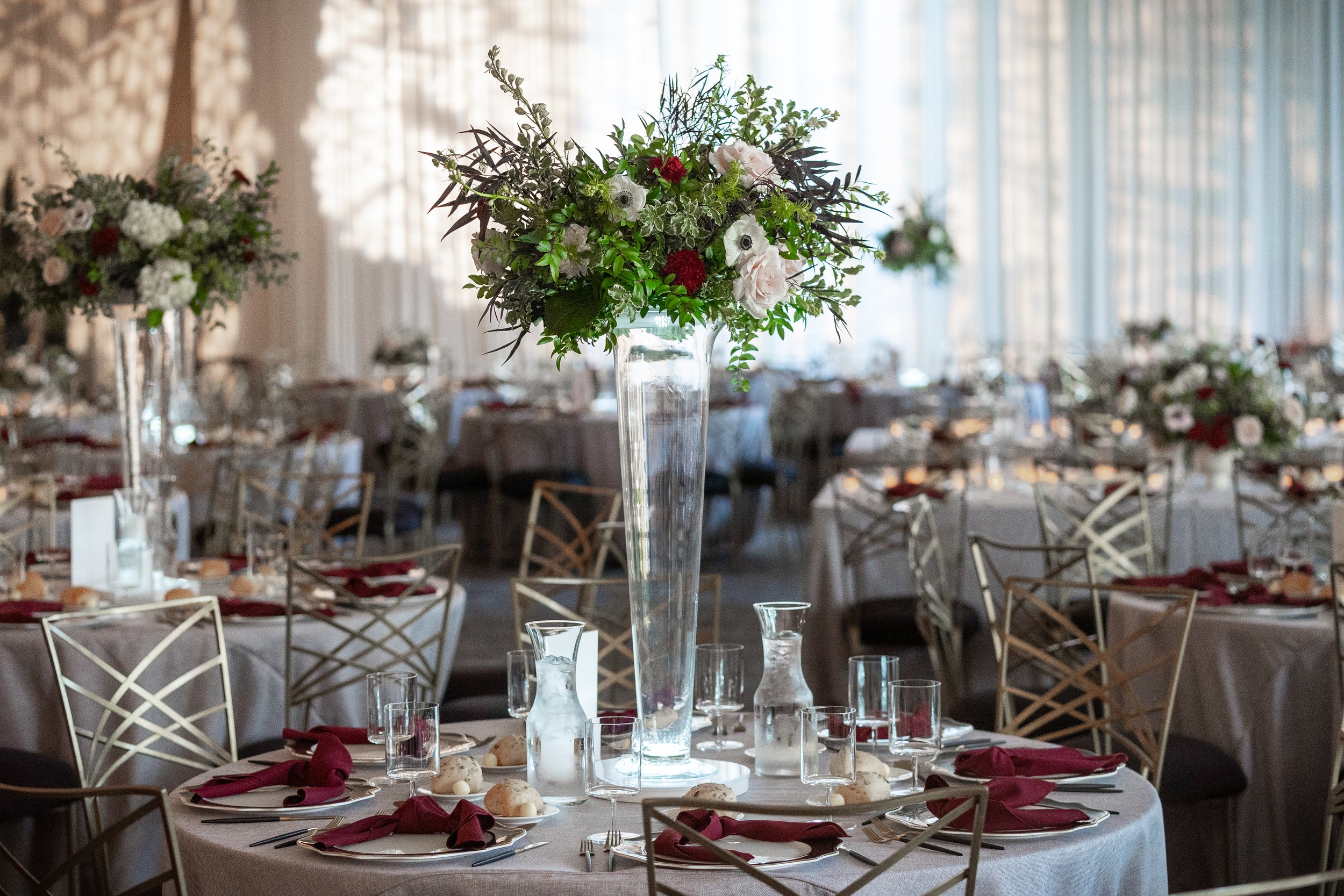 Elegant banquet hall decorated with tall centerpieces of flowers on round tables set for a formal event.