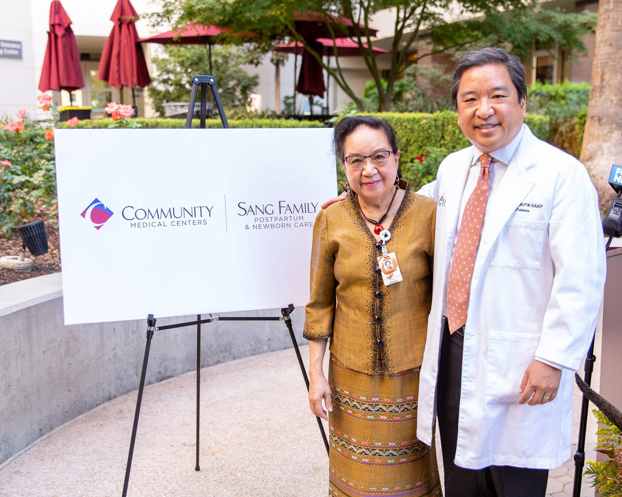 A woman and a man standing outdoors next to a sign that reads 'Community Medical Centers' and 'Sang Family Postpartum & Newborn Care.' The woman is wearing a traditional dress and the man is wearing a white medical coat and tie, smiling and posing fo