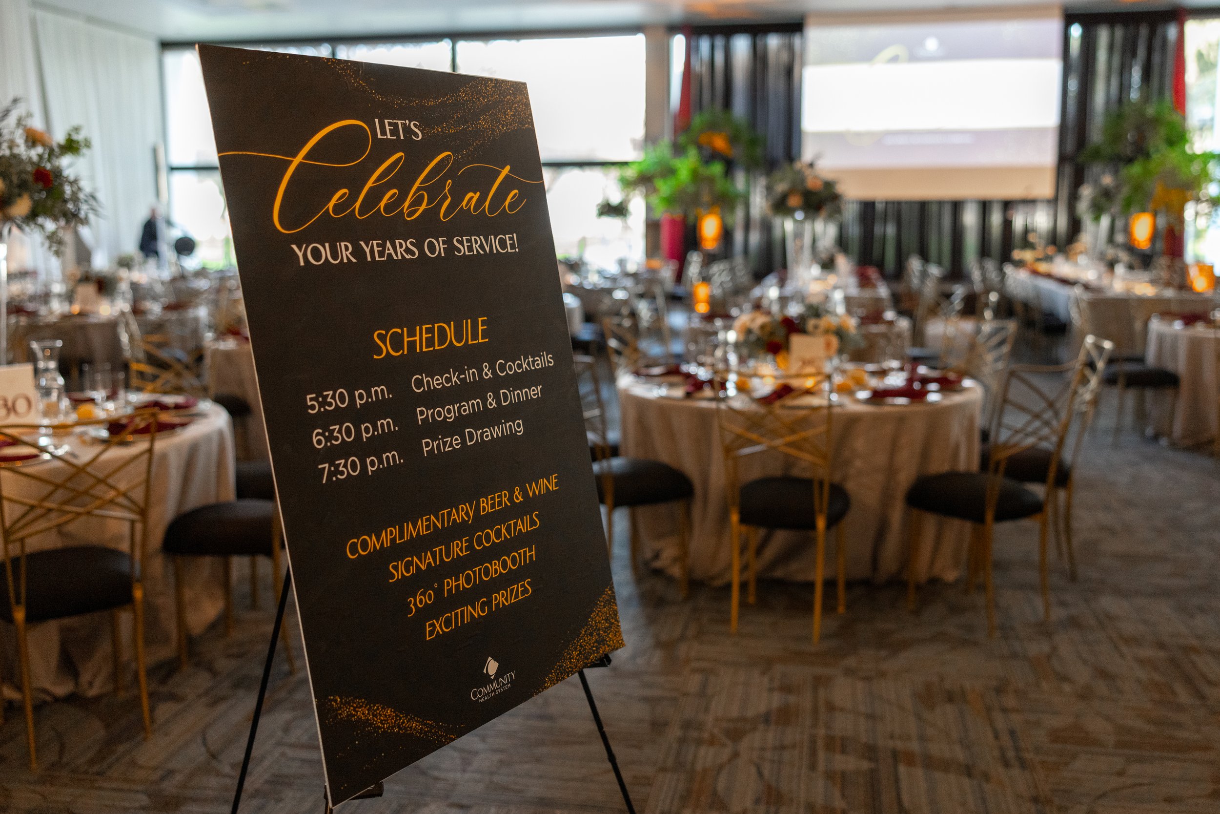Event schedule sign at a celebration banquet with round tables decorated with centerpieces, chairs, and a large screen in the background.