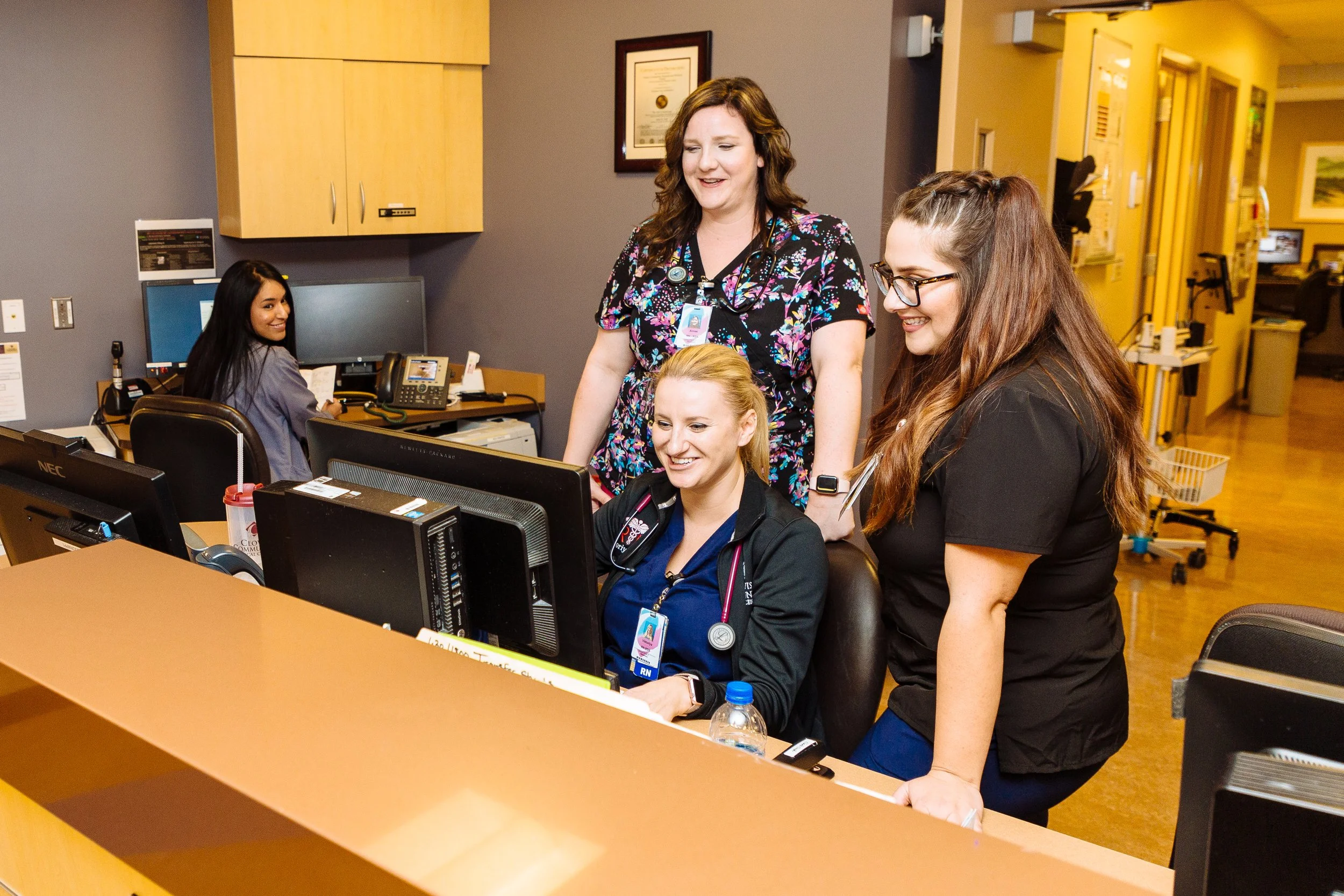 Three healthcare professionals in scrubs and uniforms working together at a computer in a hospital or clinic, with a patient in the background.
