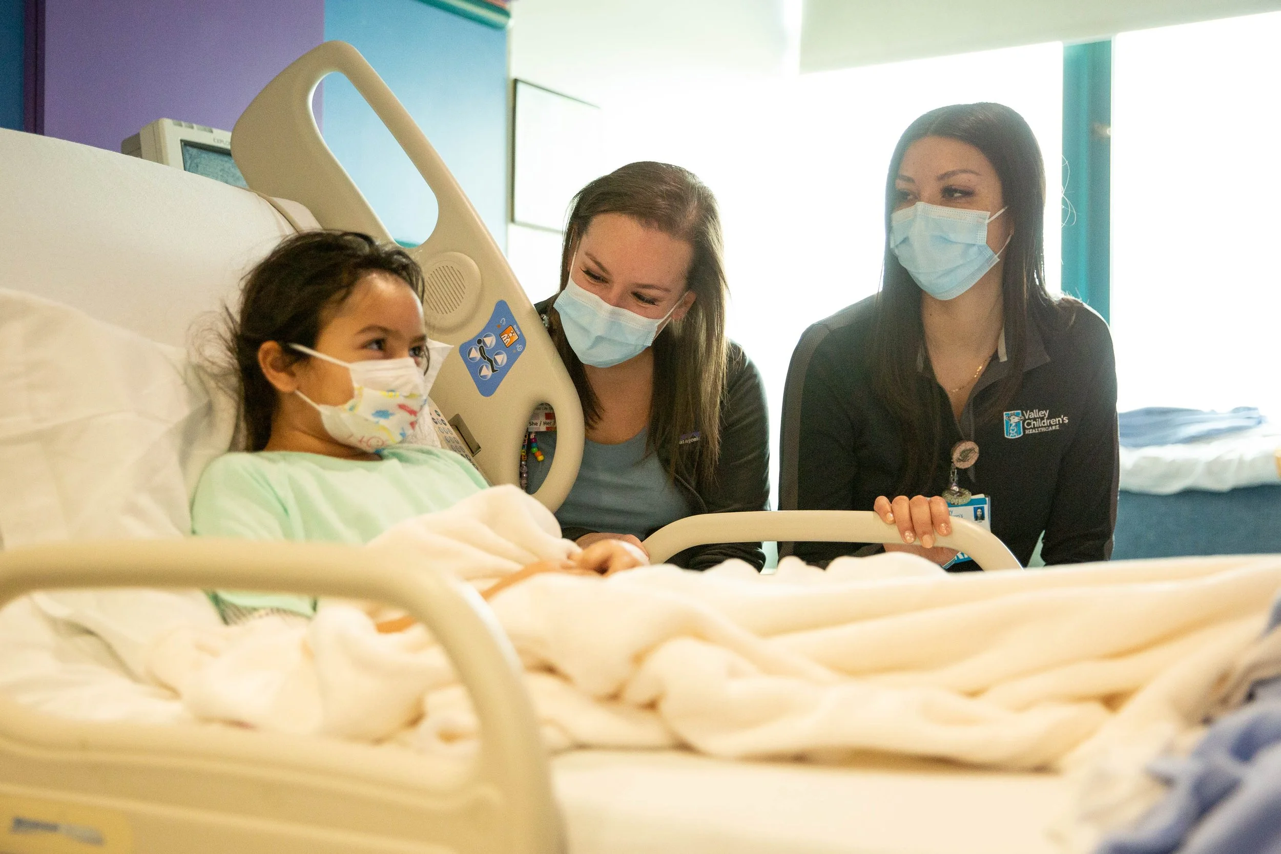 A young girl in a hospital bed wearing a face mask, with two hospital staff members visiting her, all wearing face masks in a brightly lit hospital room.