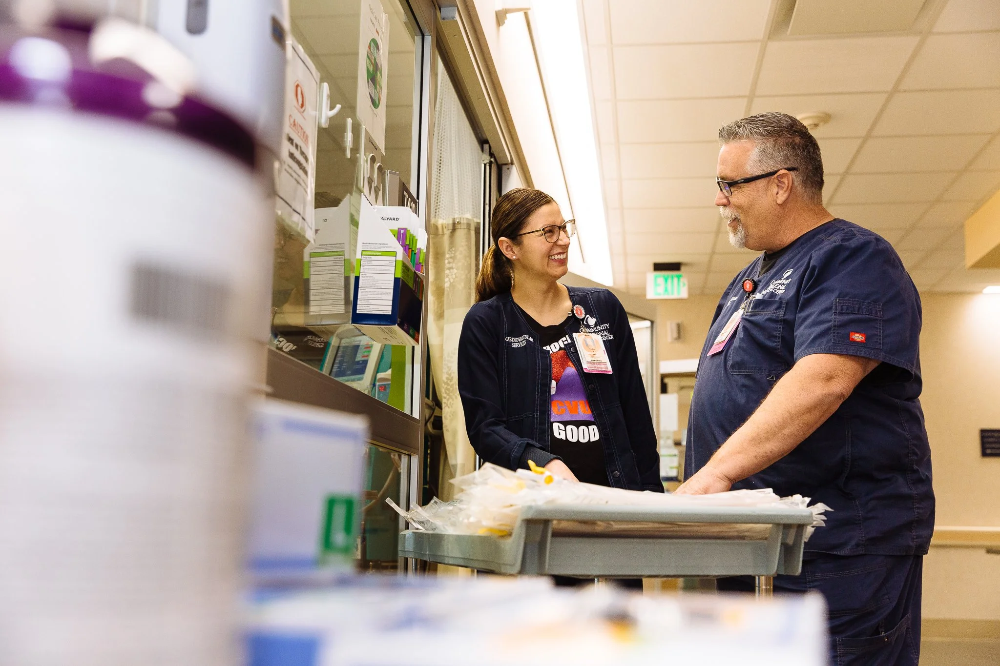 Two healthcare workers, a woman and a man, are smiling and talking to each other in a hospital setting. The woman is wearing glasses and a black shirt with a purple and orange logo, while the man has gray hair and is wearing scrubs. They are standing
